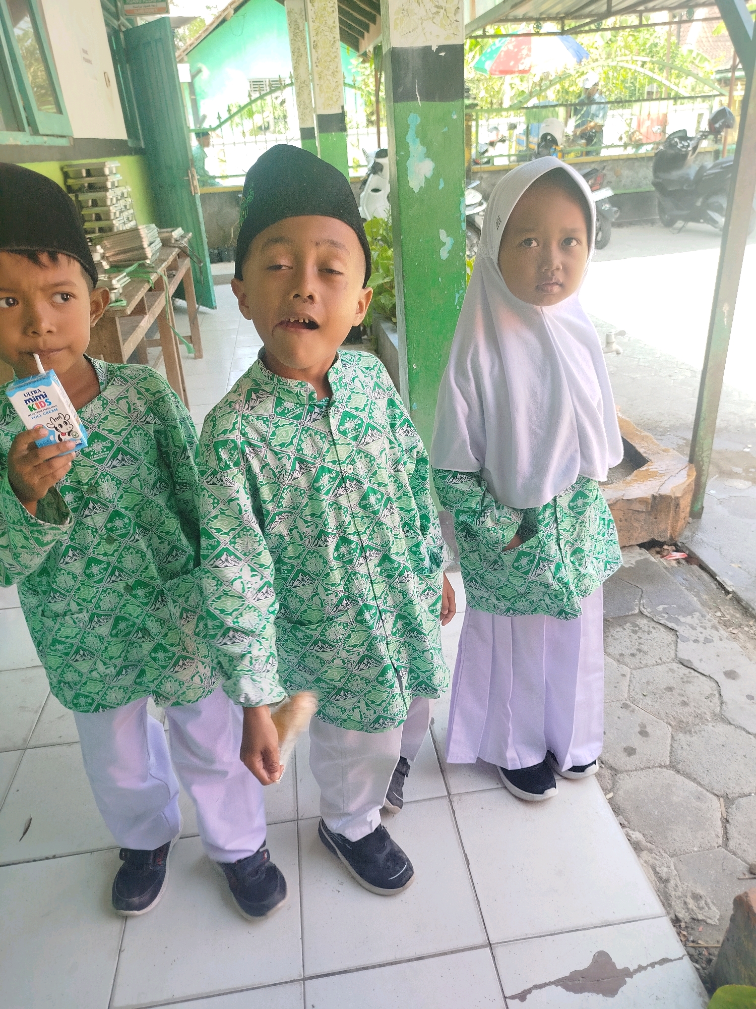 Three children are standing in front of a building in what appears to be a schoolyard in Semin, Indonesia. The child on the left, a boy wearing a black skullcap and a green and white patterned shirt, is holding a carton of milk and drinking from a straw. The boy next to him, also wearing a black skullcap and a matching green and white patterned shirt, is looking at the camera with his mouth open. To the right of the boys stands a younger girl wearing a white hijab and a long white skirt, her face also directed towards the camera. They are all wearing white pants. In the background, there is a covered walkway with a railing, some motorcycles, and vegetation. The ground is tiled in places and paved with hexagonal stones in others.