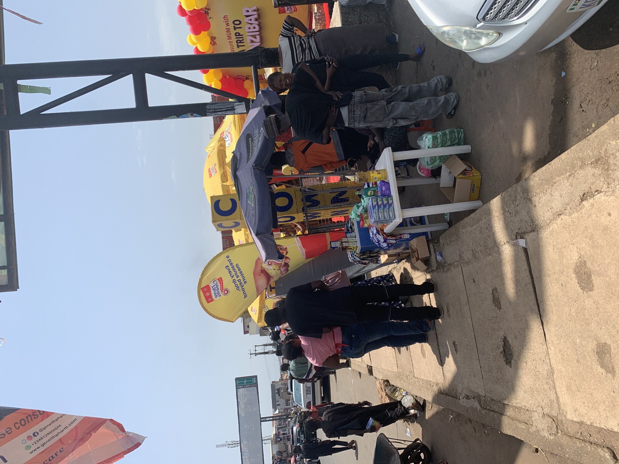 The image depicts a street scene in Jos, Nigeria, featuring a busy market stall under a sunny sky. Several individuals are present, including vendors and shoppers. A prominent yellow banner advertises a "TREASURE MUM with TRIP TO ZANZIBAR" promotion. Various products, including bottled drinks and packaged goods, are displayed on tables and shelves. The background shows a street with passing vehicles and buildings, indicating an urban setting. The sidewalk is made of concrete slabs, and there is evidence of some debris on the ground. The overall atmosphere suggests a lively commercial area with active trade.