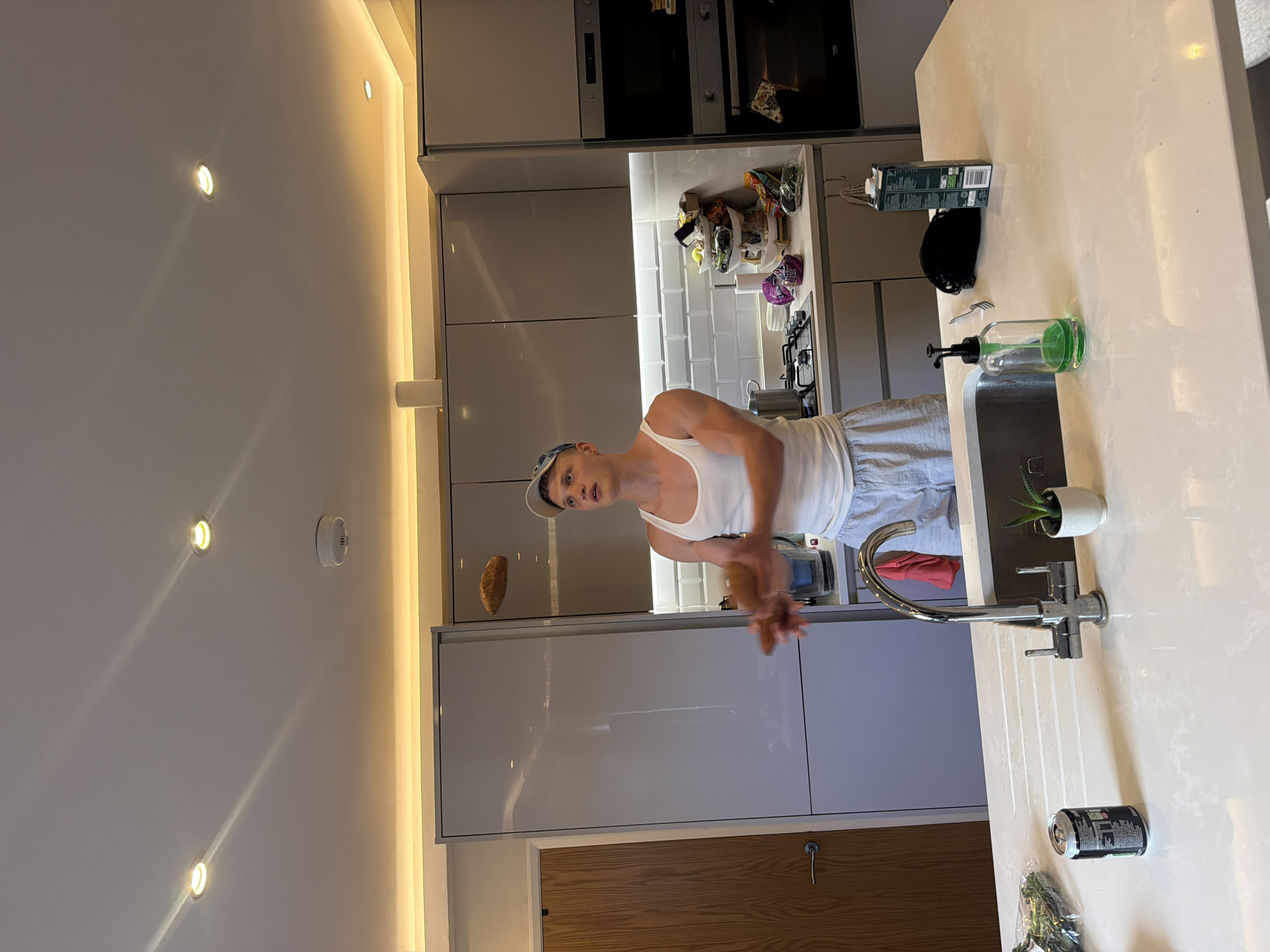 A man is in a modern kitchen. The kitchen is brightly lit by overhead spotlights. The man is wearing a white tank top and a cap. He is facing the camera, looking surprised, and is gesturing with his hands. To his left, there is a potato floating in the air. Behind him are kitchen cabinets and shelves with food and other items on them. In front of him, there is a kitchen counter with a sink, a faucet, a bottle of soap, and a plant in a pot. There are also other items on the counter, such as a can and some greens. The location appears to be inside a residential building, based on the appearance of the interior design and the lack of outdoor scenery. It is not possible to definitively determine if this location is in the city of London, United Kingdom.