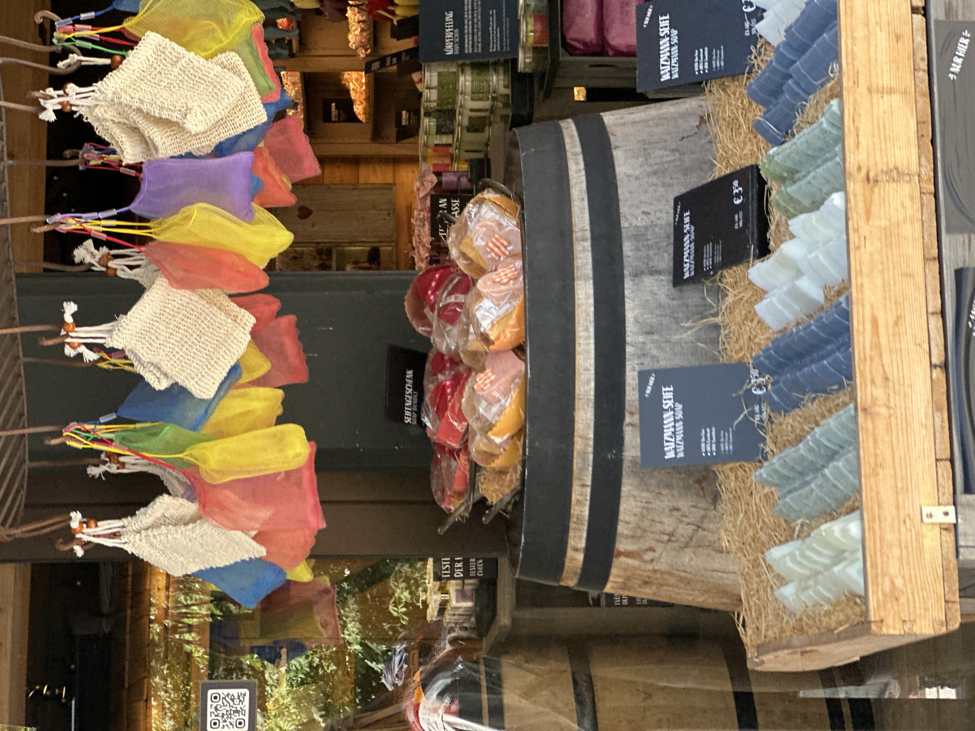 The image depicts a retail display in Schönau am Königssee, Germany, featuring artisanal soaps. In the foreground, a wooden crate is filled with rows of intricately shaped soaps, resembling mountain peaks. These soaps are arranged on a bed of straw, with light blue, light green, and dark blue hues visible. Two black display cards are placed behind the soaps, one with text including "WATZMANN-SEIFE" and "WATZMANN-SOAP," and a price of "€3.50" is visible on another card.
Behind the crate, a large wooden barrel with black bands serves as a riser for further display items. On top of the barrel, several red, orange, and pink items are packaged in transparent plastic, possibly more soaps or bath products. These are arranged on a tiered stand.
Further back, hanging from a wooden structure, are colorful mesh bags filled with what appear to be soap pouches, in shades of red, yellow, green, blue, and purple. A QR code is visible on a glass surface to the left of the hanging bags.
The background reveals a store interior with shelves stocked with various jars and containers, suggesting a retail environment selling bath and body products. The overall scene conveys a rustic and natural aesthetic, likely promoting local or handcrafted goods.