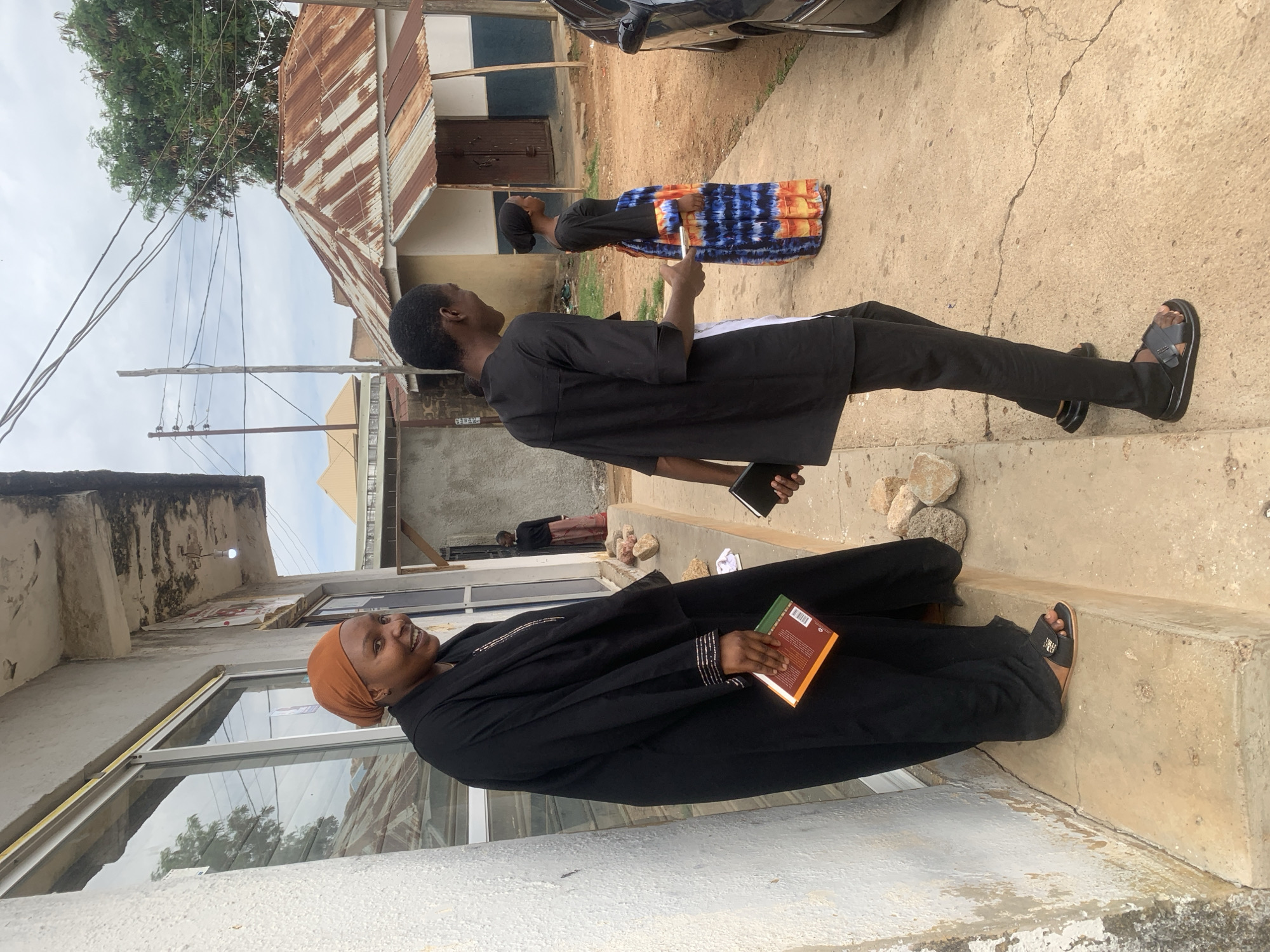 The image depicts an urban street scene in the city of Jos, Nigeria, featuring four individuals and multiple structures under an overcast sky.
In the foreground on the left, an adult female is positioned on a concrete step, facing partially right and smiling towards the camera. She wears a black abaya with silver detailing on the sleeves and collar, an orange head covering, and black open-toed sandals. She holds a small orange and green book in her right hand. Behind her is a white-painted concrete building with large, framed glass windows, above which a bare light bulb hangs from a weathered overhang.
To the right, an adult male walks away from the camera, looking towards the right side of the frame. He wears a black top with a white side panel, black trousers, and black open-toed sandals. He holds a black smartphone in his left hand and a smaller black object, possibly a notebook, in his right. He is stepping onto a concrete ground surface that shows cracks and dirt.
Further in the background on the right, another adult female stands, facing left. She wears a black head covering, a black long-sleeved top, and a long multi-colored tie-dye skirt featuring blue, orange, and yellow patterns. Adjacent to her is a small, single-story building with a severely rusted corrugated metal roof and a wooden door. The building's walls are partially painted light yellow and dark blue.
In the far background, a fourth individual, an adult female in a dark top and patterned skirt, is partially visible standing in a doorway to the left of the small building. Overhead, a wooden utility pole supports numerous tangled electrical or communication wires. Green trees are visible behind the buildings. A portion of a dark-colored car is parked on the dirt ground on the far right. The overall environment suggests an established, utilitarian urban area.