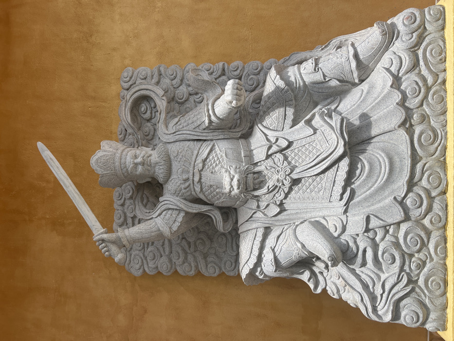 A grey stone statue of a male figure is depicted against a textured, plain yellow-orange wall in Đăk Mar, Vietnam. The figure, possibly a deity or warrior, wears an ornate crown and elaborate, patterned armor, including a prominent dragon head embellishment on the chest. It possesses a full beard, mustache, and a stern facial expression. The statue is rendered in a seated or poised posture, with its right arm raised high, gripping a carved stone sword pointing upwards and to the left. The left hand is clenched into a fist. The figure is flanked by and rests upon intricately carved cloud-like patterns, which form part of its base and surround its form. Shadows suggest outdoor lighting conditions.
