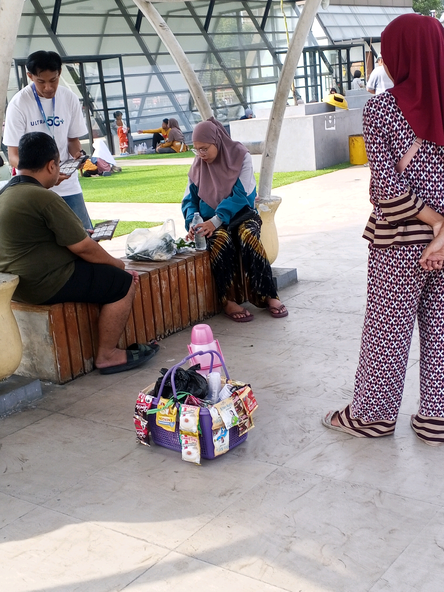 In Serang, Indonesia, at a covered outdoor area with a modern, angular roof structure, three individuals are present. A man wearing a white t-shirt with "ULTRA 5G+" branding and jeans is standing and interacting with another man who is seated on a wooden bench. The seated man, wearing a dark green t-shirt and black shorts, is holding a tablet. Beside him on the bench are a plastic bag and a small object. A woman in a teal and brown hijab and patterned clothing is seated on the same bench, holding a plastic bottle of water and looking downwards. To the right, another woman wearing a maroon hijab and a patterned maroon and white outfit is standing and facing away from the camera. In the foreground, a purple woven basket is filled with various items, including plastic bags, food packets, and a pink water bottle. The ground is paved with light-colored square tiles, and a grassy area is visible in the background.
