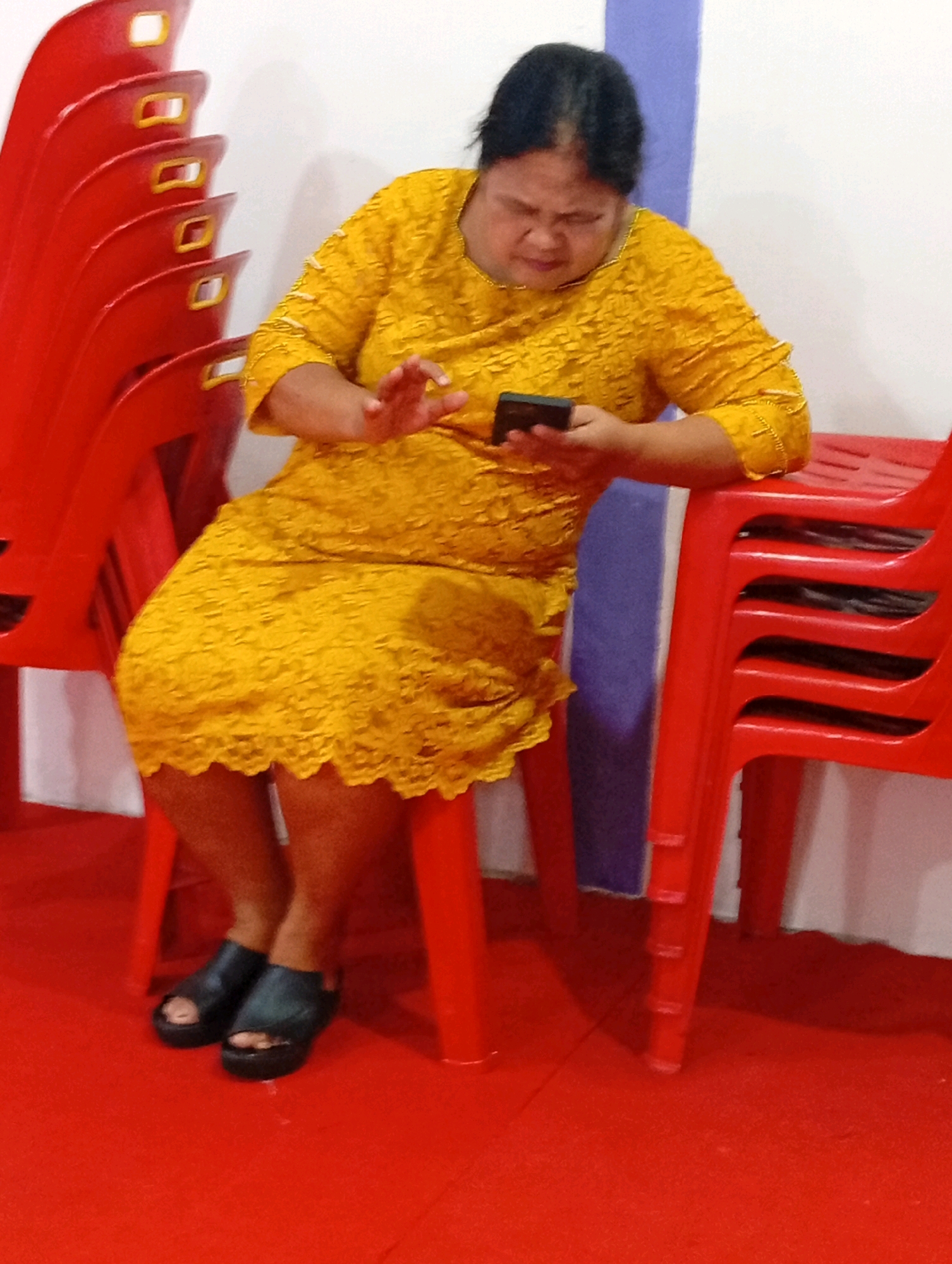 A woman wearing a yellow lace dress and black sandals is seated on a red plastic chair in what appears to be a room with a red floor covering and a white wall with a purple vertical stripe. She is looking down at a black smartphone in her hands. To her left, a stack of red plastic chairs is visible.