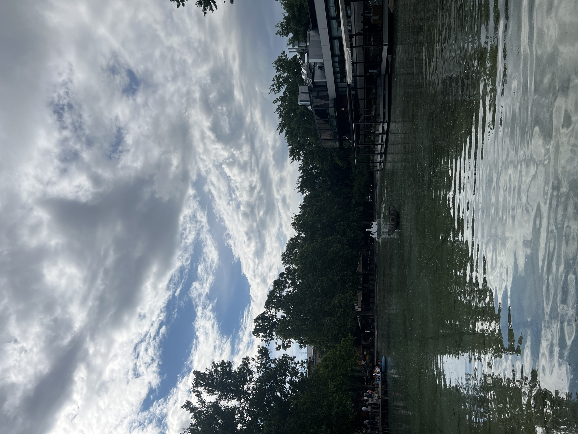 The image shows a body of water, possibly a canal or river, with a building on the right side and trees lining the bank. The sky is filled with dramatic clouds. There appear to be people near the water's edge. The presence of a fountain in the water adds to the scene. The location is Yerevan, Armenia.