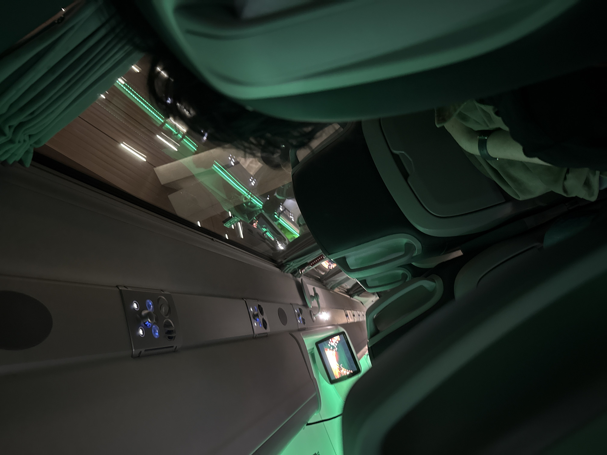 The image shows the interior of a train carriage. The interior is modern and well-lit, with white walls and dark green accents. There are several rows of seats visible, with monitors on the backs. The overhead luggage compartments are closed. The outside is visible through the window, featuring city lights and buildings. Based on the overall environment and style, this is a train that operates in Varna, Bulgaria. There is a person seated in the carriage.