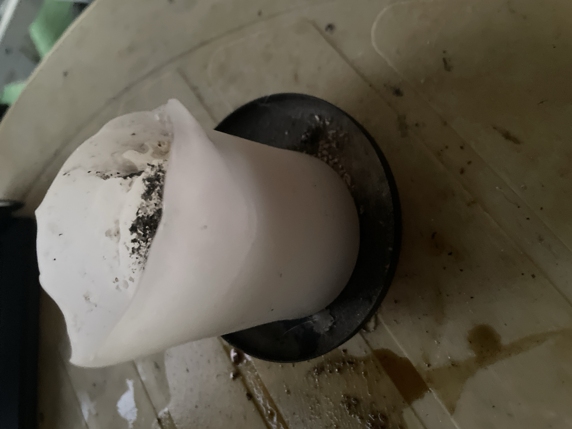 A partially melted white candle sits in a small black candle holder on a wooden surface. The candle is unevenly melted, with some blackened ash and debris on its top. The wooden surface is stained with dark liquid, possibly wax or another substance, and appears to be outdoors. The overall scene suggests a neglected or abandoned setting.