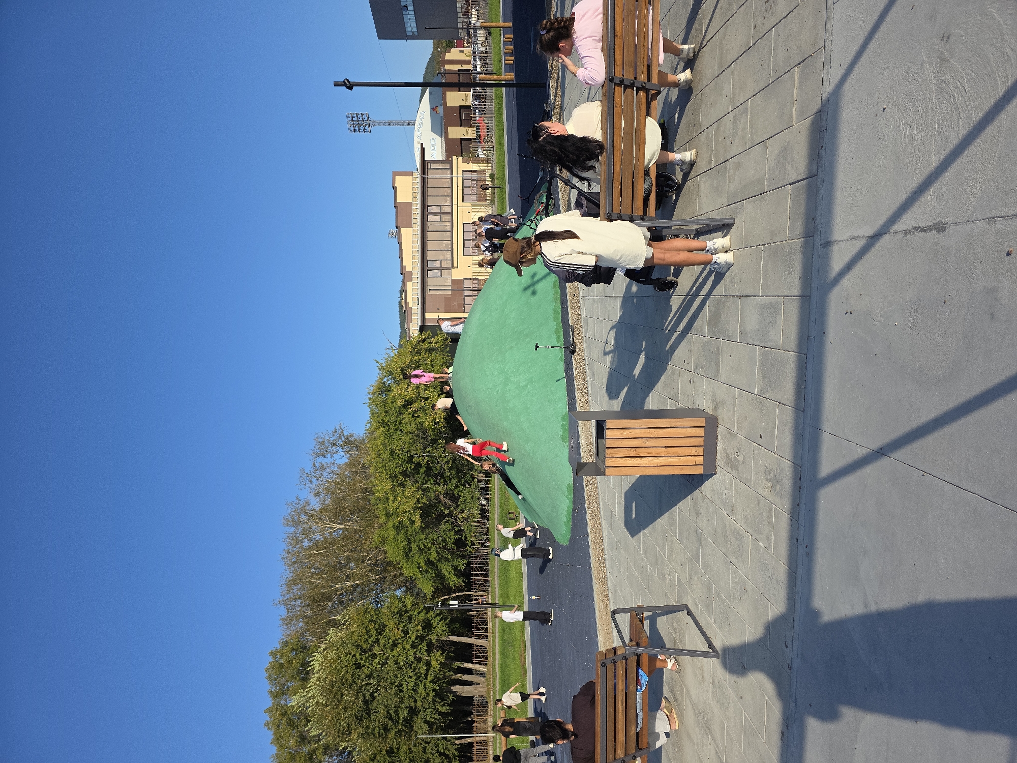 The image depicts a modern children's playground with a distinctive green, dome-shaped climbing structure. Several children are actively engaged on and around this structure, with one child in red pants ascending the slope and another in a pink outfit near the summit. The playground area features a blue rubberized surface and is surrounded by a paved area with benches.
In the foreground, three individuals are seated on a wooden bench, facing away from the camera. Two of them appear to be young girls, one wearing a pink top, and the third person might be an adult. Further to the left, another person is seated on a bench, facing the playground.
Behind the playground, a line of trees forms a natural backdrop, beyond which are modern buildings, including a multi-story structure with numerous windows and a stadium with floodlights. The sky above is a clear, vibrant blue, indicating fair weather. The overall scene suggests a public recreational space within a city.
The image is likely taken in Miass, Russia, given the request. While the specific location within Miass cannot be determined from the visual information alone, the presence of modern urban infrastructure and a well-maintained public space suggests it could be a newly developed park or recreational area. The architecture of the buildings in the background may offer clues to those familiar with the city's landmarks.