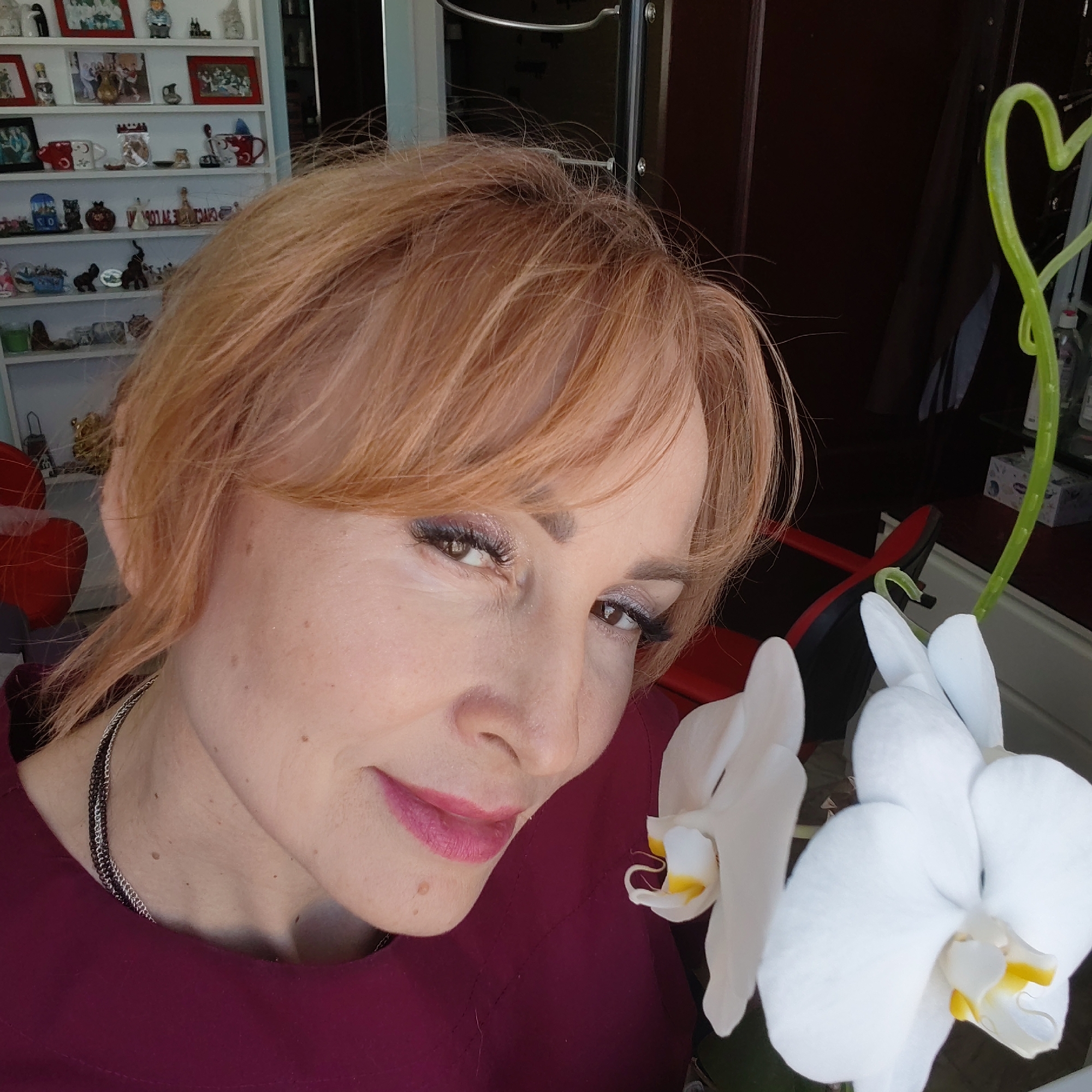 A woman with reddish-blonde hair, wearing a maroon top and a layered silver necklace, is photographed in what appears to be an indoor setting. She has makeup on, including eyeshadow and lipstick. A white orchid is in the foreground. In the background, there is a shelf with various figurines and decorative items. To the right of the woman is a dark-colored door or cabinet. The location is Chaikovsky, Russia.