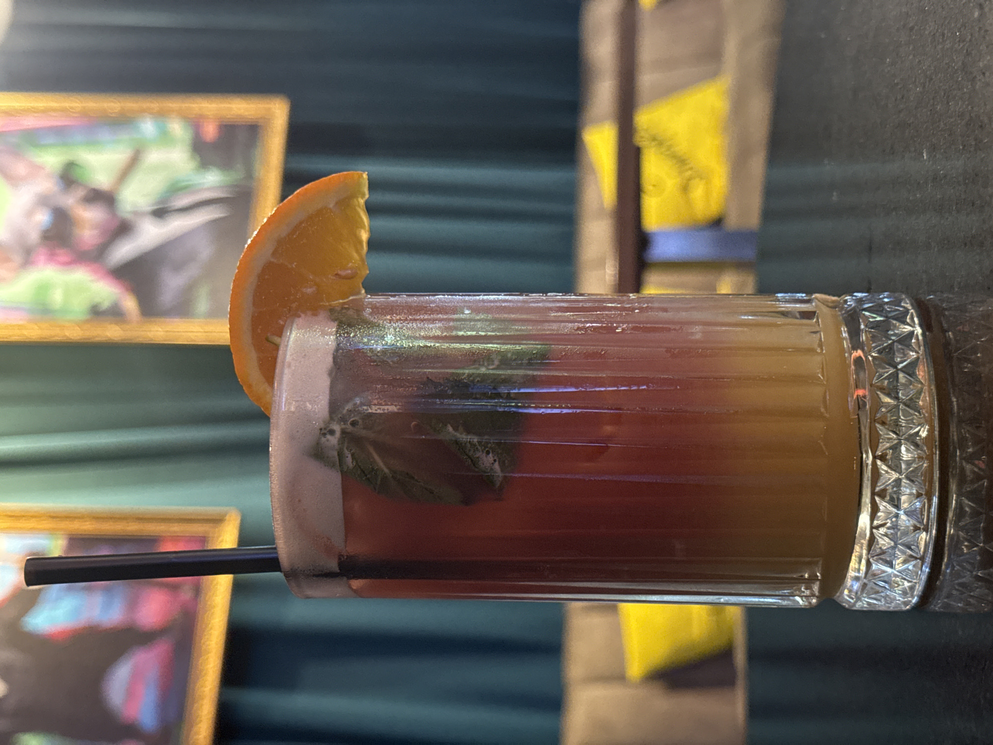 A layered cocktail with red and orange liquids, mint leaves, and an orange slice garnish sits in a fluted glass on a dark, reflective surface, likely a bar or table. A black straw is inserted into the drink. In the background, teal curtains hang behind framed artwork. Two framed paintings are visible: one on the left with abstract colors and a dark figure, and another on the right depicting a stylized animal wearing a suit and sunglasses, holding a cigar. Cushioned seating with yellow pillows is visible in the blurred background, suggesting an indoor establishment. This scene is in Varna, Bulgaria, at a venue called "Bar Zareni Gradishi".