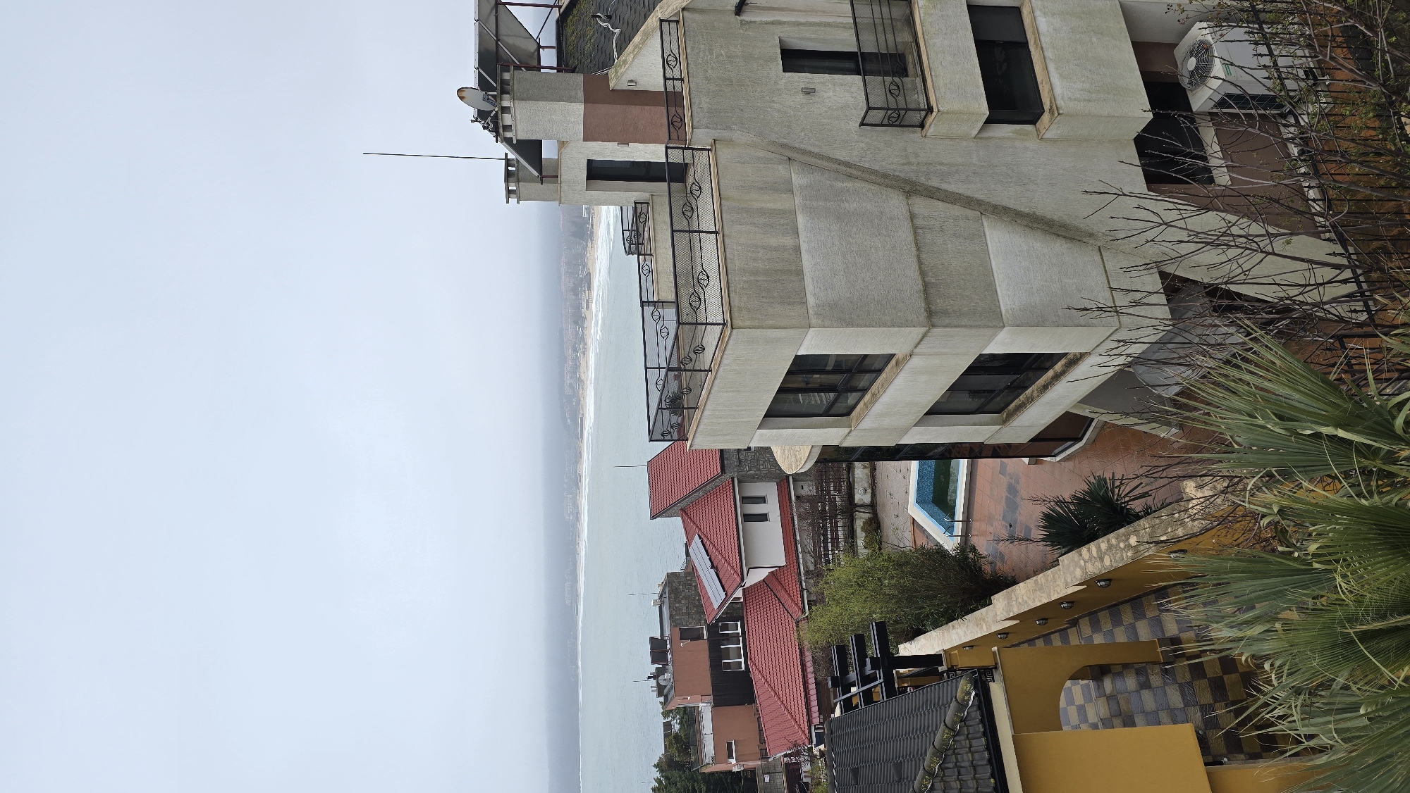 The image presents an elevated view of Sozopol, Bulgaria, with a focus on a multistory building and the cityscape extending toward the sea. The sky is overcast, creating a subdued atmosphere.
The primary building is of modern architectural design, featuring a concrete-like facade with multiple balconies and windows. Details such as railings and satellite dishes on the roof are visible. Adjacent to this building are other residences with red tile roofs, suggesting a residential area. A blue swimming pool is also visible, adding to the residential context.
The sea stretches into the distance, with a visible shoreline. The overall environment suggests a coastal urban landscape in Sozopol. The image is devoid of people, focusing instead on the built and natural elements of the scene.