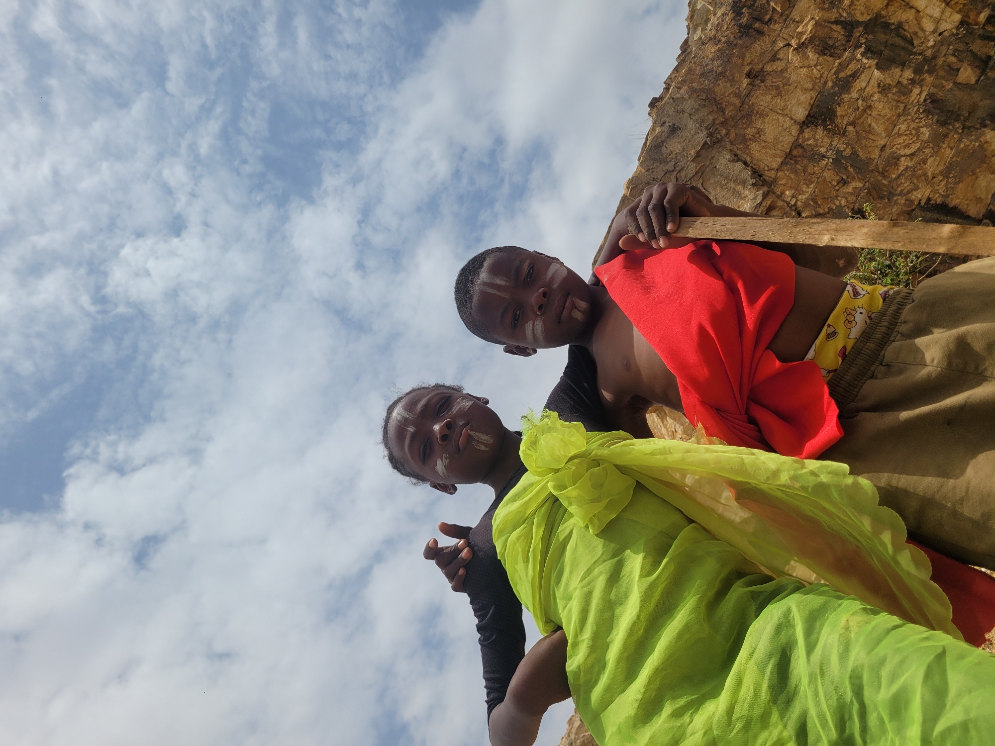Two children, with markings on their faces, are positioned in the foreground against a backdrop of a partially visible, weathered rock formation and a sky filled with clouds. The camera's perspective is from below, looking up at the subjects. The child on the right wears a red cloth draped over them, while the child on the left is draped in a bright green cloth. The scene takes place in what is most likely the city of Barakin Akawo, Nigeria. One child holds a wooden stick.