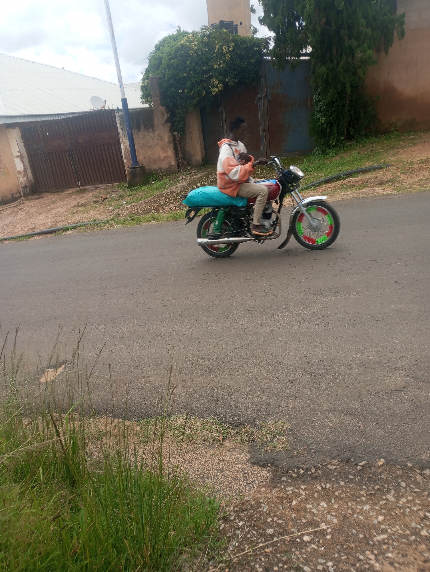 In Jos, Nigeria, a person is riding a motorcycle on a paved road. The person is wearing a light-colored hoodie with orange accents and khaki pants, and they are seated on the motorcycle which has a turquoise-colored cover over the rear seat. The motorcycle has red and green painted spokes on its front wheel and appears to be carrying a light blue bag secured on the back. The environment shows a roadside with overgrown green grass and some dirt. In the background, there is a brown gate, a building with a white corrugated roof, and a tall tree with dark green foliage. The sky is overcast with clouds.