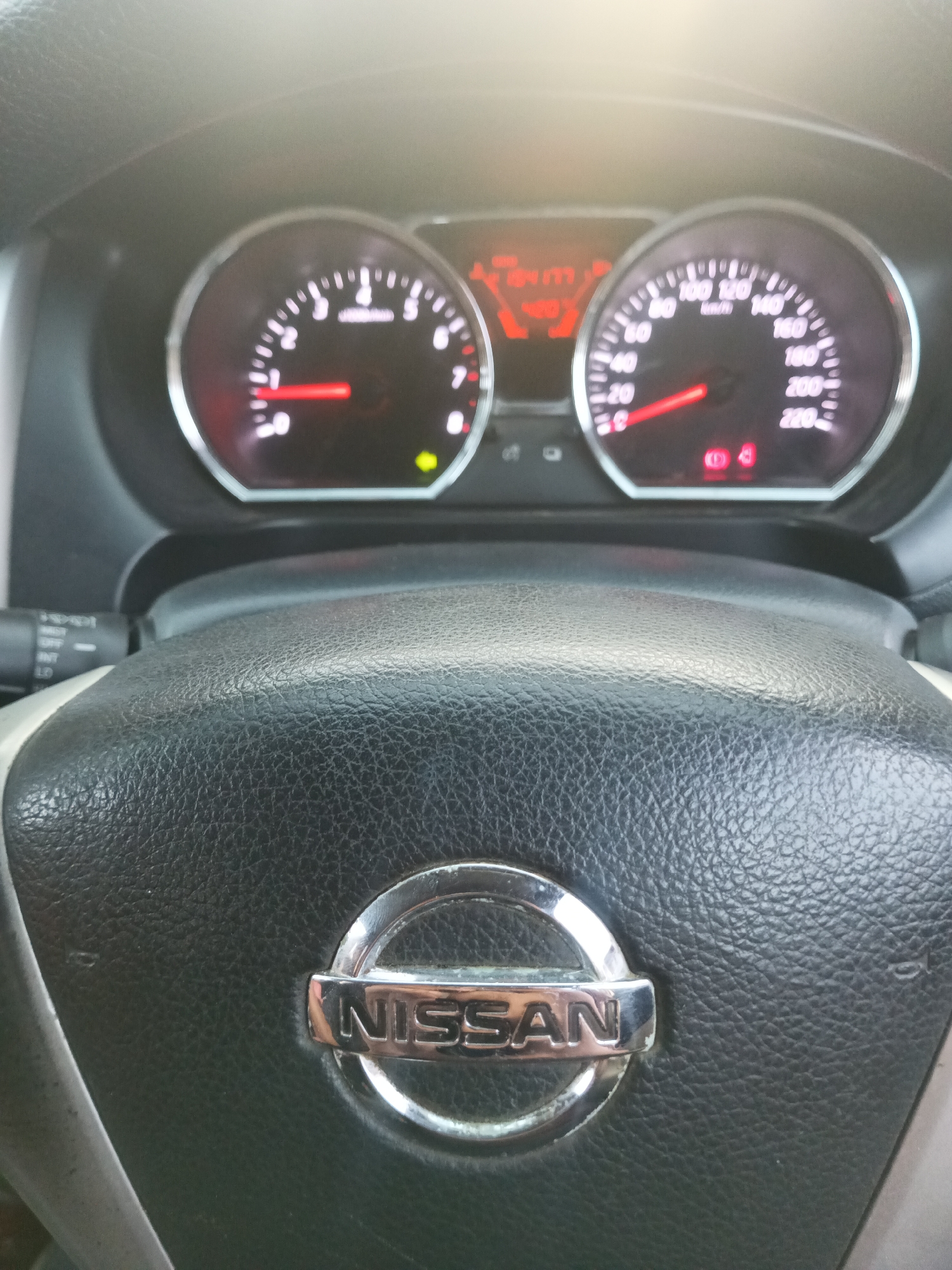 The image shows the interior of a Nissan vehicle, focusing on the steering wheel and dashboard. The steering wheel is black and textured, with a prominent silver Nissan logo in the center. To the left of the steering wheel, a turn signal lever is visible. The dashboard features two circular gauges: a tachometer on the left and a speedometer on the right. Both gauges have red needles and white markings. The tachometer indicates a reading between 0 and 1, and the speedometer shows a reading of approximately 100 km/h. There are also various indicator lights visible on the dashboard, including a red symbol that appears to be an airbag warning light and a green arrow indicating turn signals. The overall lighting suggests it is daytime, with some bright glare from the sun visible at the top of the image. The vehicle appears to be parked or in motion in an environment that is not discernable from the image itself.