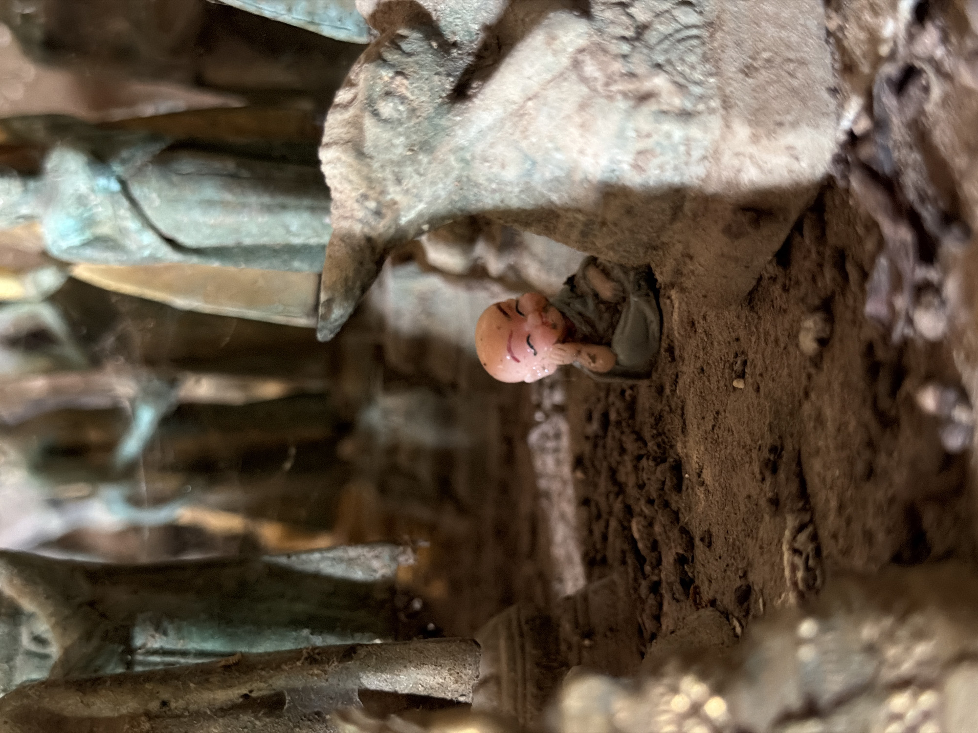 A shallow depth of field shot shows a miniature clay figurine of a monk sitting in the foreground. The monk is depicted with closed eyes, a smile, and one hand resting on its cheek. It is wearing a simple grey robe. Behind the monk, there are larger, weathered statues of figures, partially obscured by a rough, textured surface that appears to be made of rock or stone. The environment is earthy and appears to be a display or diorama, possibly with artificial elements. The overall lighting is dim, with some highlights on the statues and the monk. The location in Muangkeo, Laos, is not discernible from the visual elements of the image.