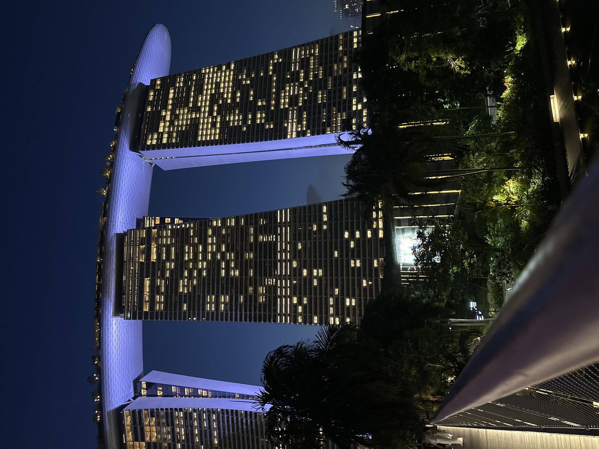 The image captures the Marina Bay Sands hotel in Singapore at night. The iconic structure, composed of three 55-story towers topped by a 340-meter-long skypark, is illuminated by a cool blue light on its upper levels. The windows of the hotel towers are lit with a warm yellow glow, suggesting occupied rooms. In the foreground, a purple railing and metal mesh fence suggest the viewpoint is from a walkway or bridge. Lush green palm trees and other foliage are visible in the middle ground, with some areas illuminated by warm lighting. In the background, the dark sky is visible, with a few distant lights from other buildings. A few figures, casually dressed, are walking on the elevated walkway on the left side of the frame. The overall scene depicts a nighttime view of a prominent Singaporean landmark, highlighting its architectural features and the surrounding urban environment.