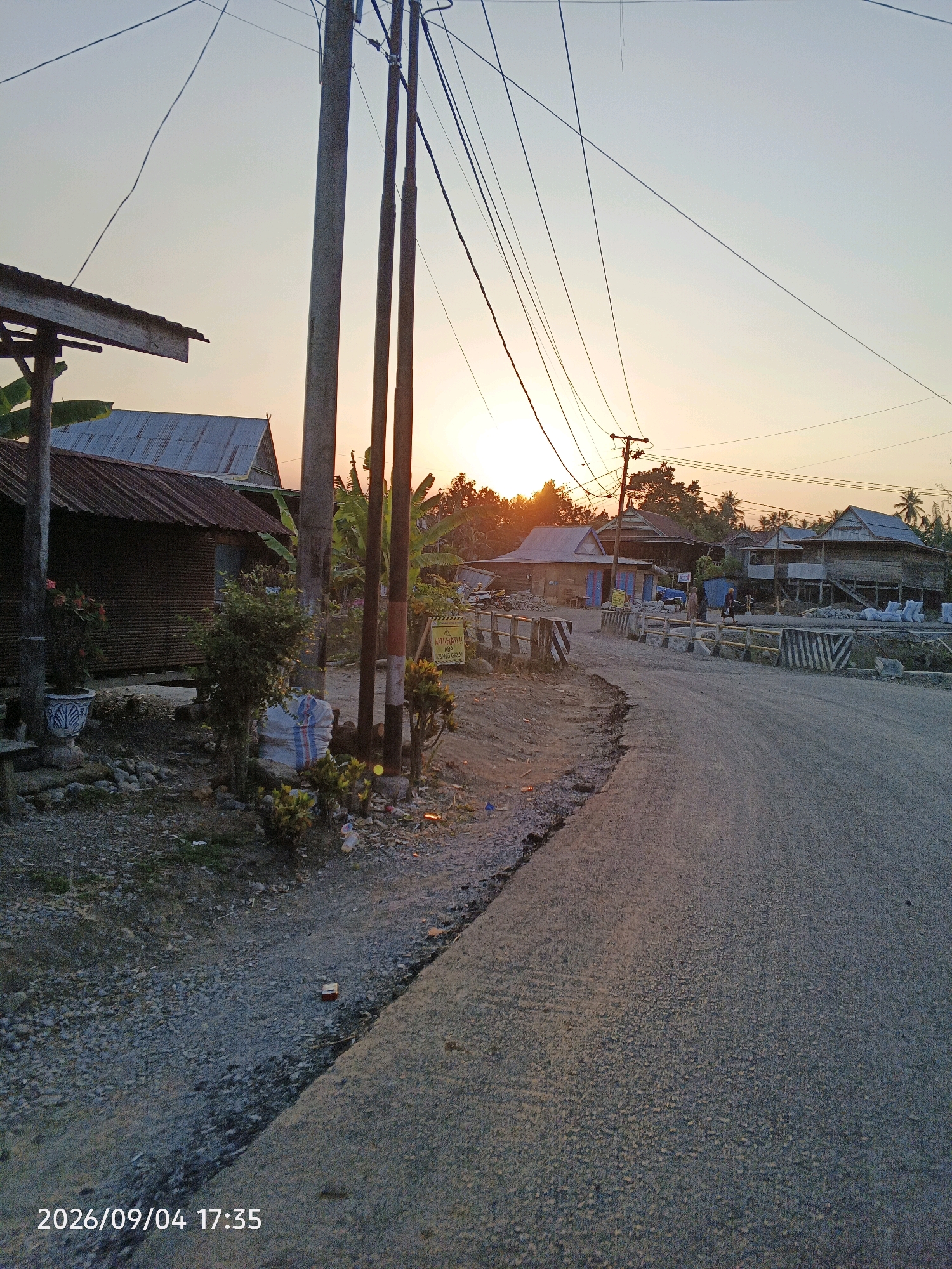 The image depicts a rural road in Bulu Cenrana, Indonesia, during sunset. The sky is a pale orange and blue, with the sun setting behind trees and buildings, casting long shadows. A dirt road curves to the right, with a rough gravel shoulder. On the left side of the road, a corrugated metal building with a tin roof stands next to a wooden structure with a canopy. Potted plants and rocks are visible near the building. Further down the road, there are more houses, some appearing to be under construction with building materials and white sacks piled up. A yellow and black striped barrier is present across a small bridge. Power lines crisscross the sky, supported by wooden poles. A yellow warning sign with black text is visible on the right side of the road, near some vegetation. The timestamp "2026/09/04 17:35" is displayed at the bottom left corner of the image. There are no people clearly visible in the scene, though two figures can be faintly discerned walking on the bridge in the distance.