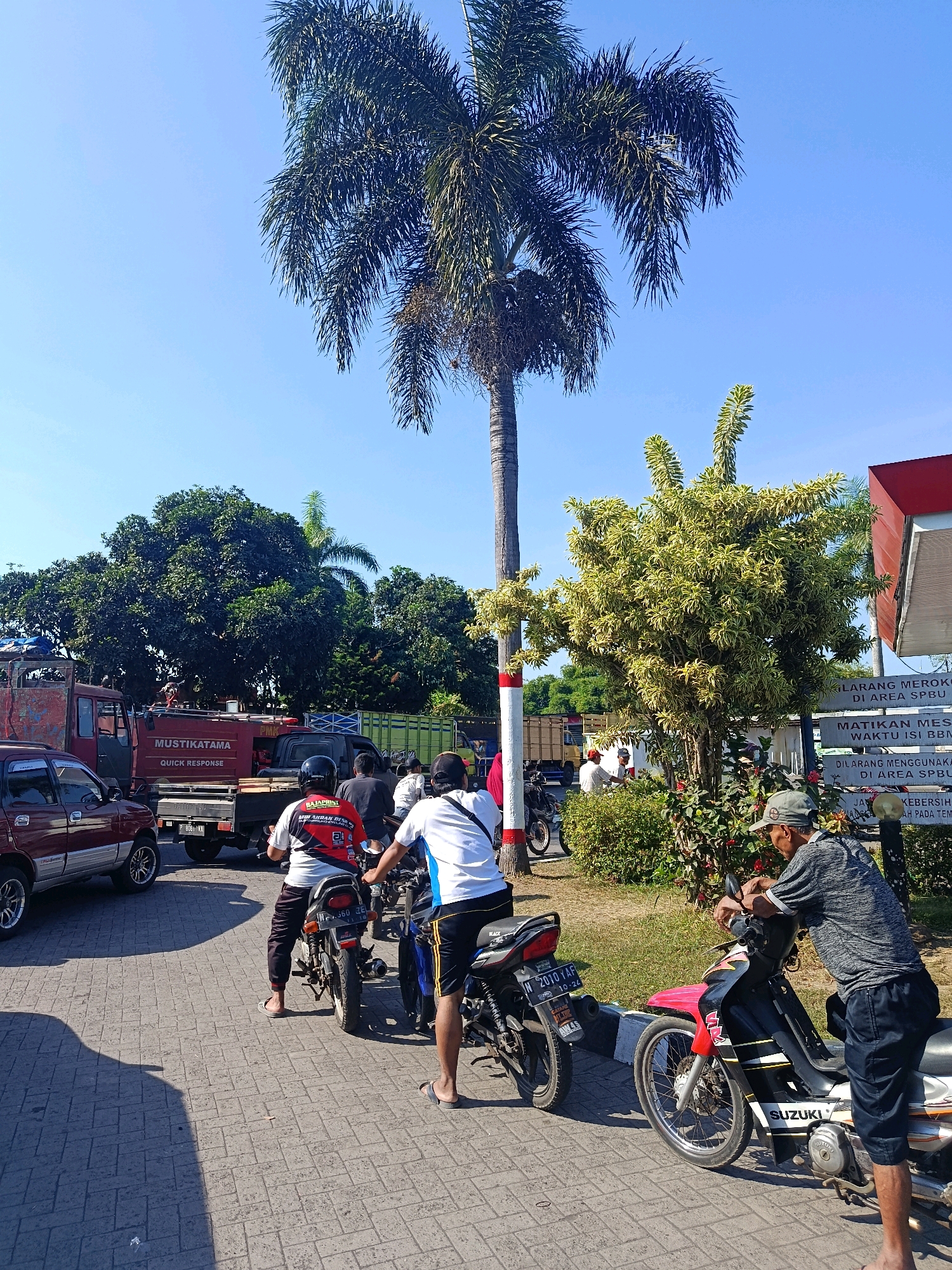The image depicts a daytime scene on a paved road in Tempeh Lor, Indonesia, likely near a gas station judging by the signage. A tall palm tree dominates the background, with a smaller, variegated tree to its right. A red truck with "MUSTIKATAMA QUICK RESPONSE" written on its side is parked on the left, with a maroon SUV in front of it. Several people are on or near motorcycles, suggesting they are either refueling or waiting in line. One man in a grey shirt and cap is leaning on a red and black Suzuki motorcycle. Another man in a white shirt with blue accents is seated on a motorcycle. A third man, wearing a black and red shirt with "BAJAPRINT" on it, is also on a motorcycle. The atmosphere appears calm, with clear blue skies overhead.