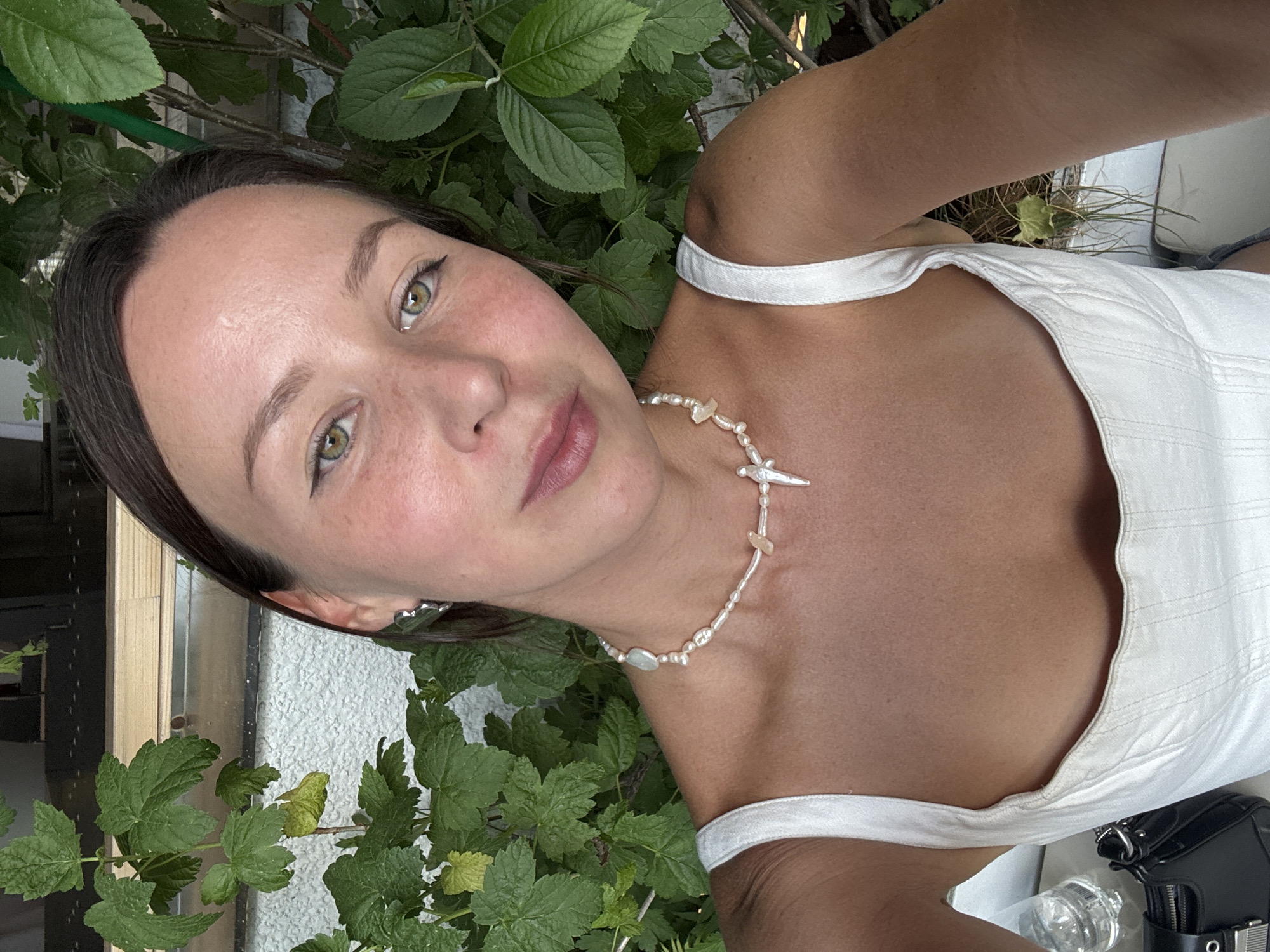 A woman is shown in a selfie, with green foliage surrounding her. She has fair skin, dark hair, and green eyes. She is wearing a white top with straps and a pearl necklace with a silver pendant. A black purse and a clear water bottle are visible in the lower right corner. The background suggests an outdoor setting near a building, possibly in a garden or courtyard. There is no information to identify the specific location in Moscow, Russia.