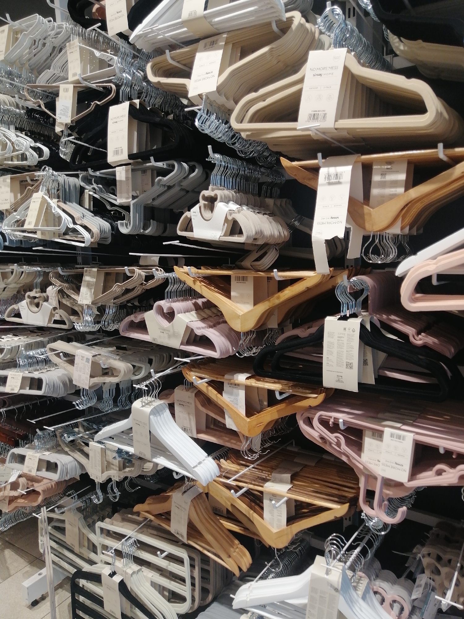 The image displays a retail store's clothing hanger display. The scene is densely packed with various types of hangers, organized on shelves and hanging racks. There are multiple rows of hangers, appearing to be made of wood, plastic, and possibly velvet, in an assortment of colors including natural wood, beige, white, grey, black, and pink. Many of the hangers have product tags attached, some with text and barcodes visible. The lighting suggests an indoor environment, likely a department store or home goods store. The overall impression is of an organized merchandise display. The location is specified as a retail store in Kremenchuk, Ukraine. There are no people visible in the image.