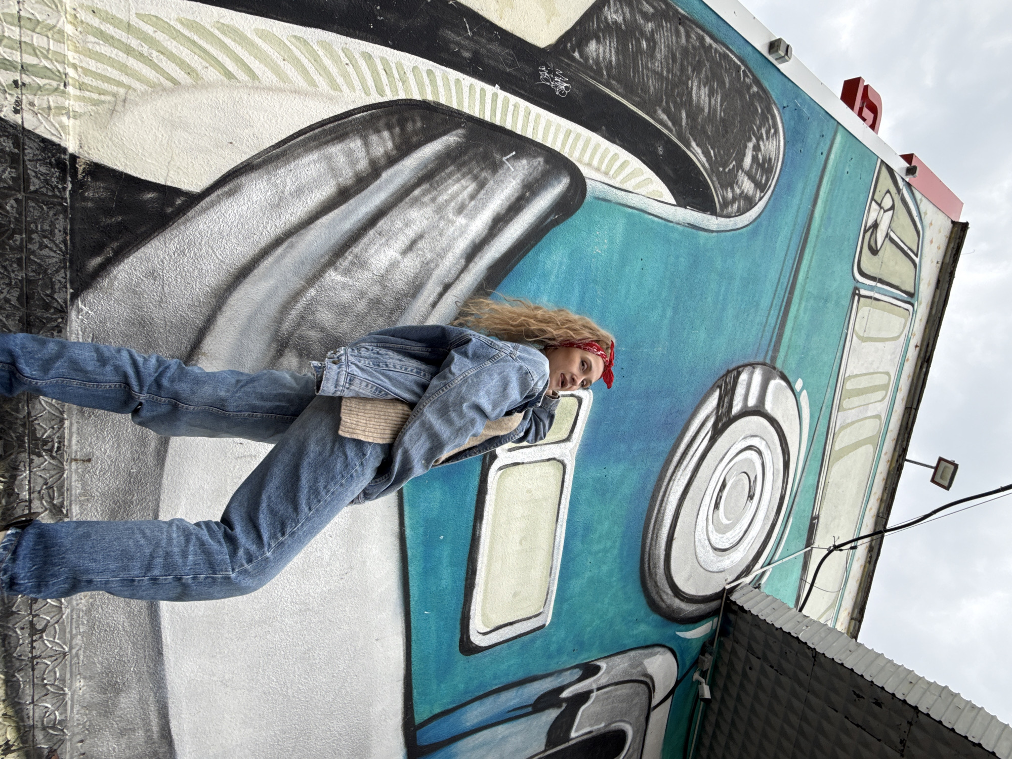 A medium shot depicts a young woman standing in front of a mural of a blue and white van on a building in the city of Chaikovsky, Russia. She is wearing a denim jacket, denim jeans, and a red bandana in her curly, blonde hair. The mural, with its illustrative style, is rendered in shades of white, grey, and blue. The building facade is visible to the right, as well as the sky above. A portion of the building's roof and a street light are also visible in the corner of the image. The woman is smiling.