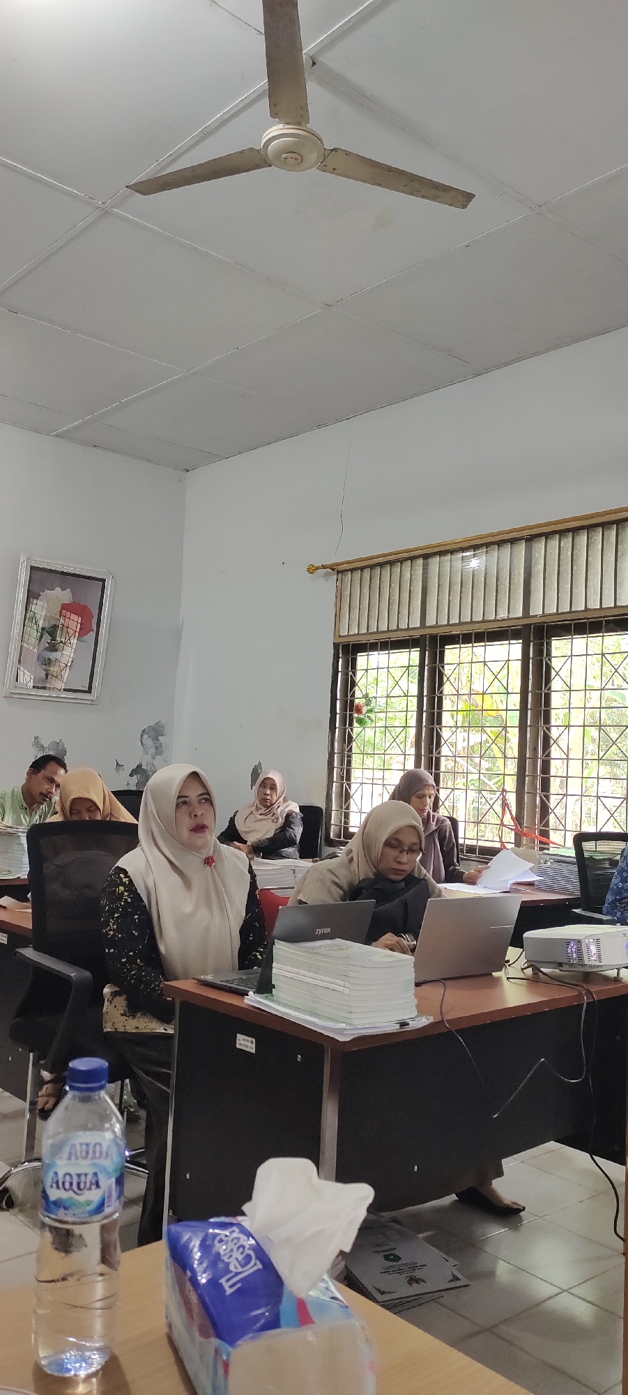 The image shows a classroom or meeting room setting. Several people, mostly women wearing headscarves, are seated at desks. Some are looking at laptops or documents, suggesting a presentation or training session is in progress. There are stacks of papers on the desks, and a projector is visible in the foreground. A water bottle and a box of tissues are also on a desk. The room has white walls, a tiled floor, and a ceiling fan. A framed picture hangs on one wall, and a window with blinds overlooks greenery. The overall impression is one of an indoor educational or professional gathering. There is no information in the image to suggest the location is the city of Langnibong, Indonesia.