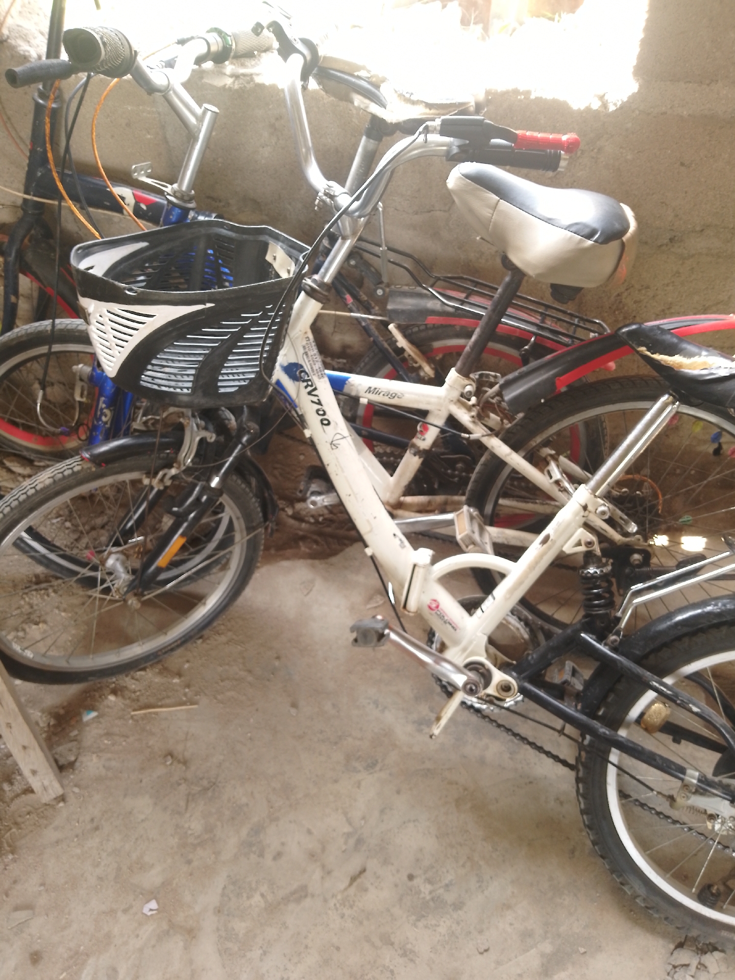 In an outdoor environment with a concrete floor and a stone wall, two bicycles are parked. The bicycle in the foreground is white with black accents, featuring a white and black basket at the front, and a red and black striped rear fender. Its rear wheel has spokes and a black tire with a white rim. The bicycle behind it is primarily black and blue with red accents on its tires and frame. The ground is littered with debris.