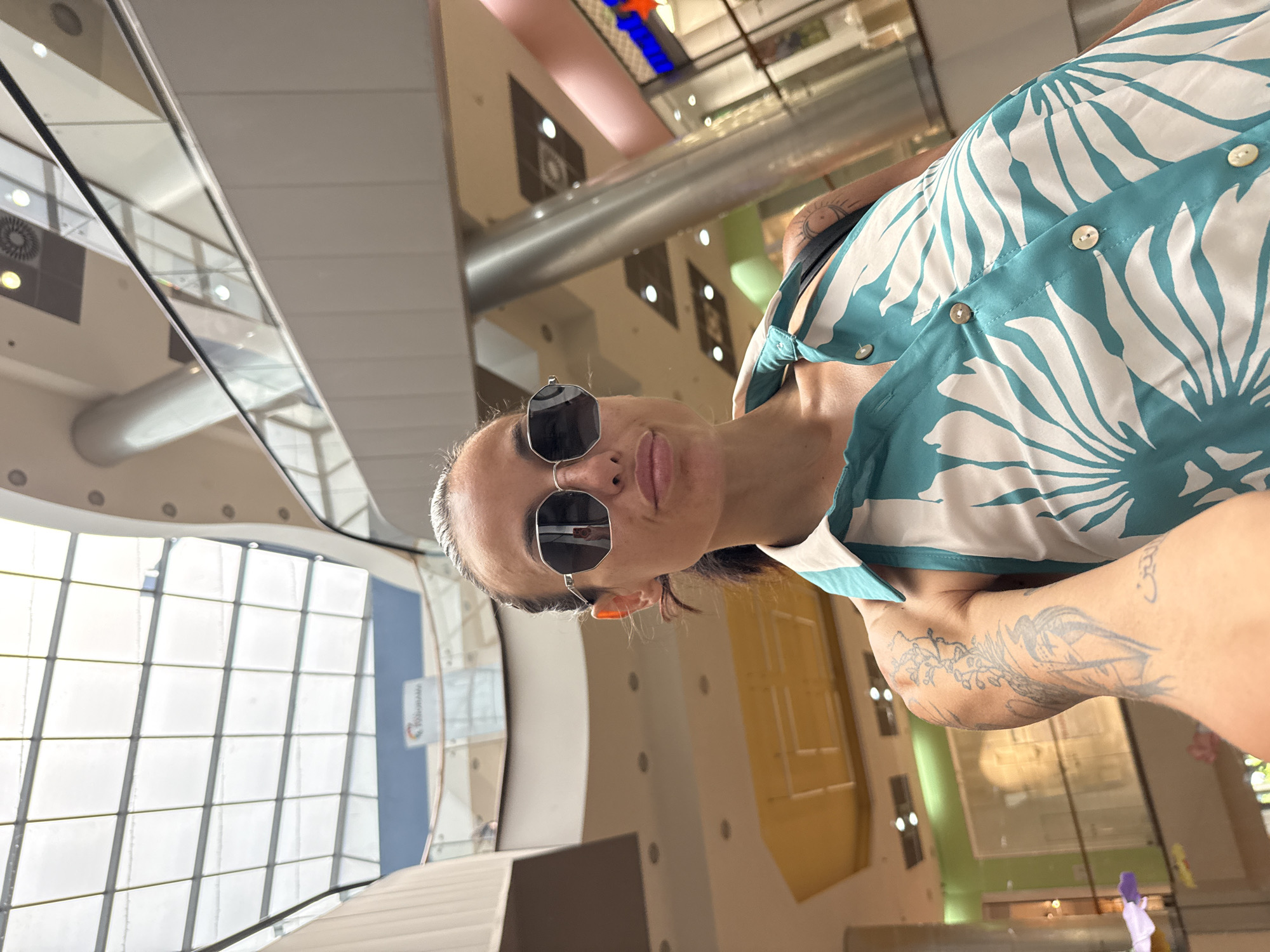 A woman is taking a selfie in a multi-level shopping mall. She is wearing sunglasses and a teal and white patterned dress. Her left arm is visible, showing tattoos. The background shows the interior of a modern mall with escalators, glass railings, and a glass ceiling. A sign with the word "PANORAMA" is visible in the distance. This is the Panorama Mall in Pleven, Bulgaria.