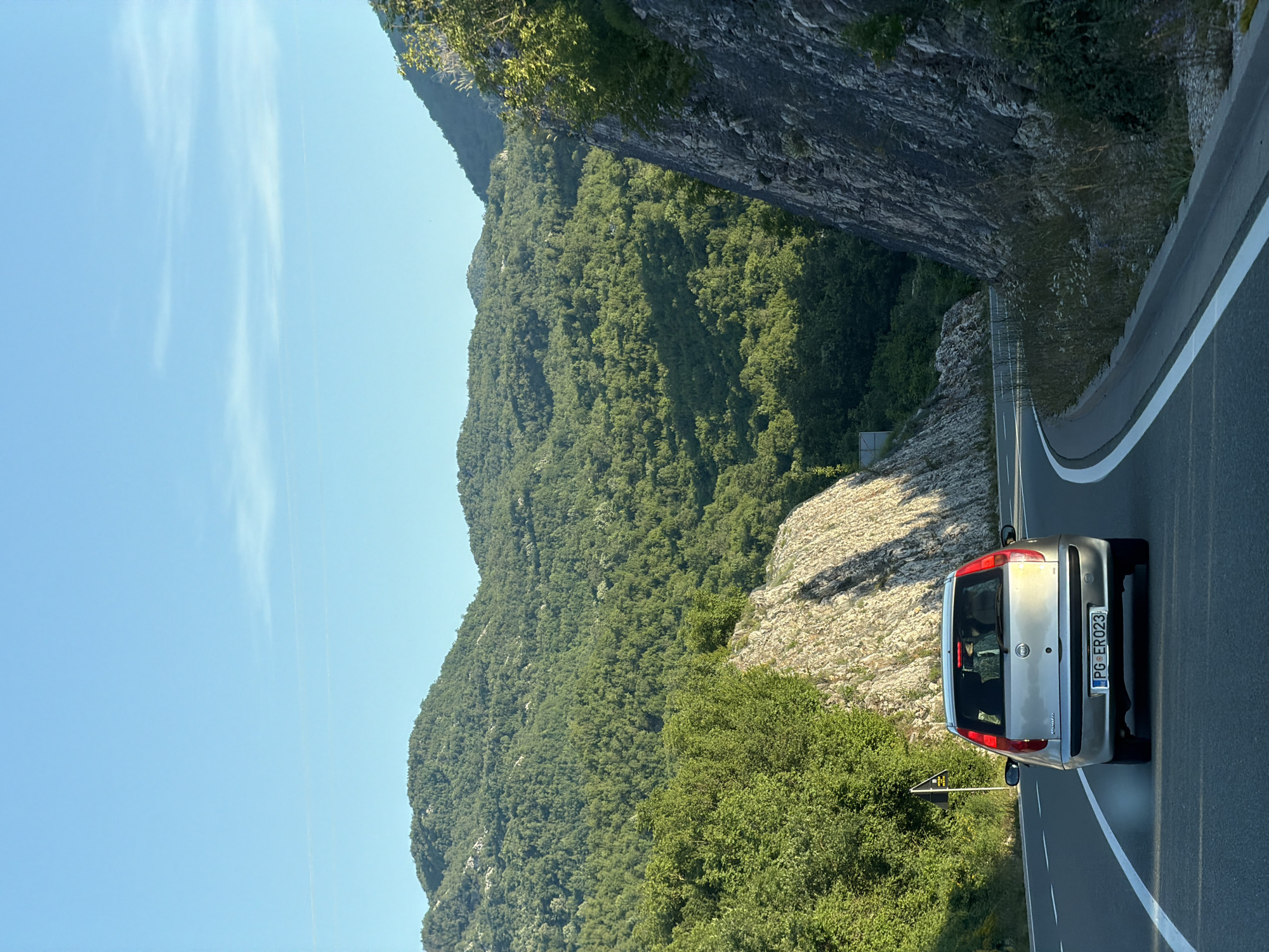 An eye-level, medium shot captures a silver Fiat Punto driving on a winding road with lush green mountains in the background. The car, seen from behind, has its brake lights illuminated. The road is asphalt with white lane markings. To the right of the road, a steep, rocky embankment is visible, covered with sparse vegetation. To the left, the terrain slopes upwards, also covered in dense greenery. The sky is clear and blue with a few wispy clouds. The scene appears to be in a mountainous area, possibly Vrela, Montenegro, given the context.