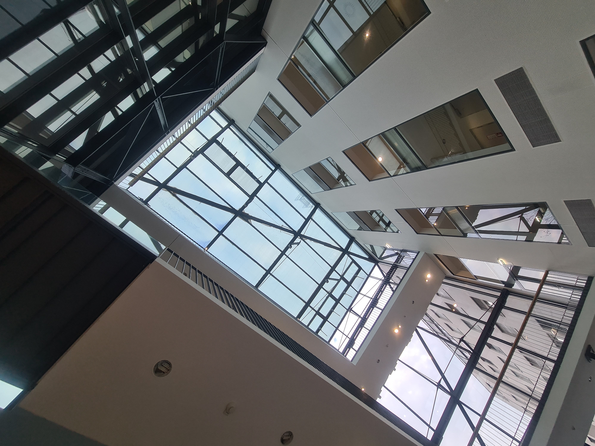 The image captures an interior view looking upwards at the atrium of a modern building, likely a corporate office or a public space with a significant architectural design. The dominant features are the extensive glass ceiling and windows, supported by dark metal framing, which create a sense of openness and allow natural light to flood the space.
The atrium has multiple levels, indicated by balconies with metal railings and illuminated walkways. The walls of the building are a light, textured material, punctuated by irregularly shaped windows that reflect the interior and exterior.
The architecture suggests a contemporary design aesthetic, with clean lines and a focus on transparency. The presence of ventilation grilles on the walls indicates a functional aspect to the building's design. The overall impression is one of spaciousness, light, and modern construction.
Without further context or visible landmarks, it is impossible to definitively pinpoint the exact location within Vienna, Austria. However, the architectural style is consistent with modern commercial or public buildings found in major European cities like Vienna.