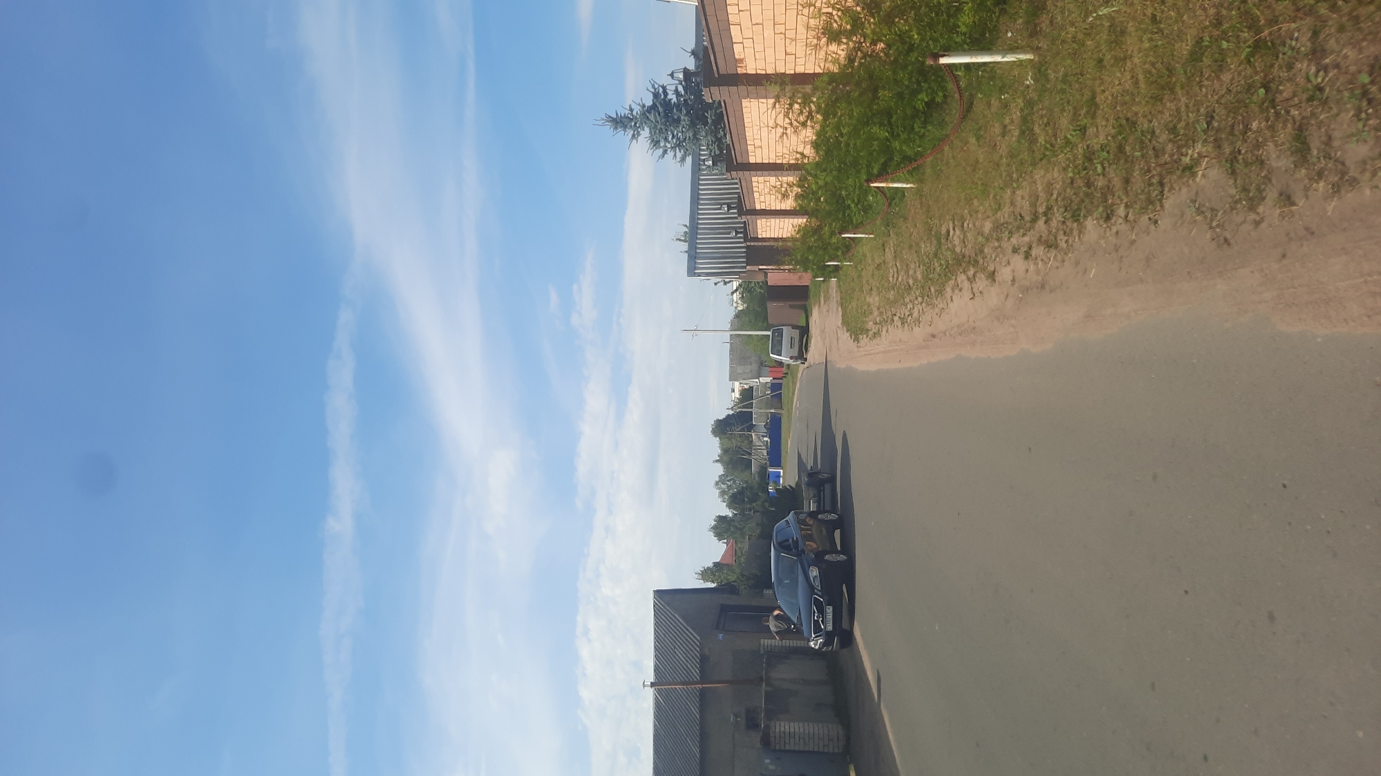 A black SUV is parked on the side of a paved road, attached to a small trailer. The vehicle is positioned in front of a grey, corrugated metal building. To the right of the road is a dirt path and a brick fence with green bushes growing along its base. In the distance, several houses and trees are visible under a blue sky with scattered clouds. The scene appears to be in a rural or suburban area.