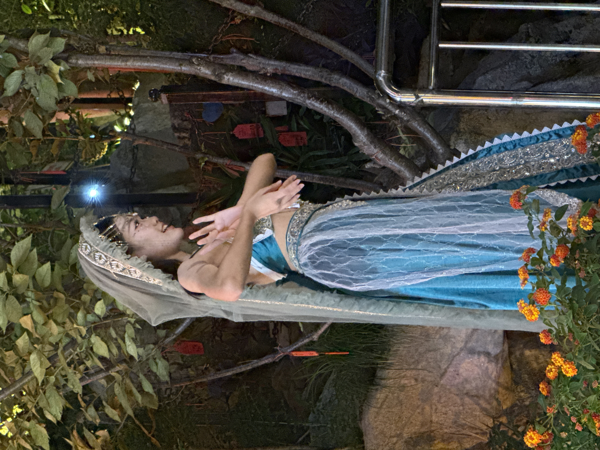 A young woman, adorned in a teal and silver costume with a translucent veil and headpiece, performs a dance amidst a naturalistic outdoor setting. The woman stands in profile, facing right, with her hands raised and her mouth open as if mid-song or exclamation. She wears a fitted, ornate bodice and a flowing skirt composed of layers of teal fabric and sheer white fabric with intricate patterns. The backdrop features a rocky embankment, gnarled trees with prominent branches, and foliage. Small red and white tags hang from some of the branches, possibly as decorations or offerings. In the foreground, a cluster of vibrant orange and yellow flowers adds a splash of color. A stainless steel railing is visible on the right side of the frame. The lighting suggests it is evening or nighttime, with artificial lights illuminating the scene. The image was captured in Yuzhong District, China.