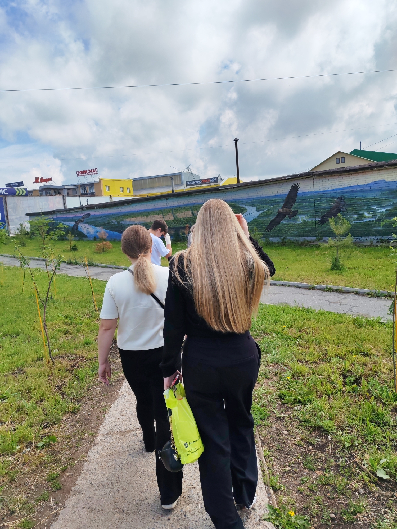 In Chaikovsky, Russia, three individuals are walking on a paved path beside a grassy area. The foremost person, viewed from behind, has long blonde hair and is wearing a black outfit. To their left, a second person with blonde hair in a ponytail wears a white top and black pants. A third person, also seen from behind, is wearing a white t-shirt and is partially obscured. Behind the individuals, a brick wall features a mural depicting birds of prey flying over a landscape with a river. Above the wall, commercial buildings with signage are visible against a cloudy sky. A bright yellow bag is being carried by one of the individuals.