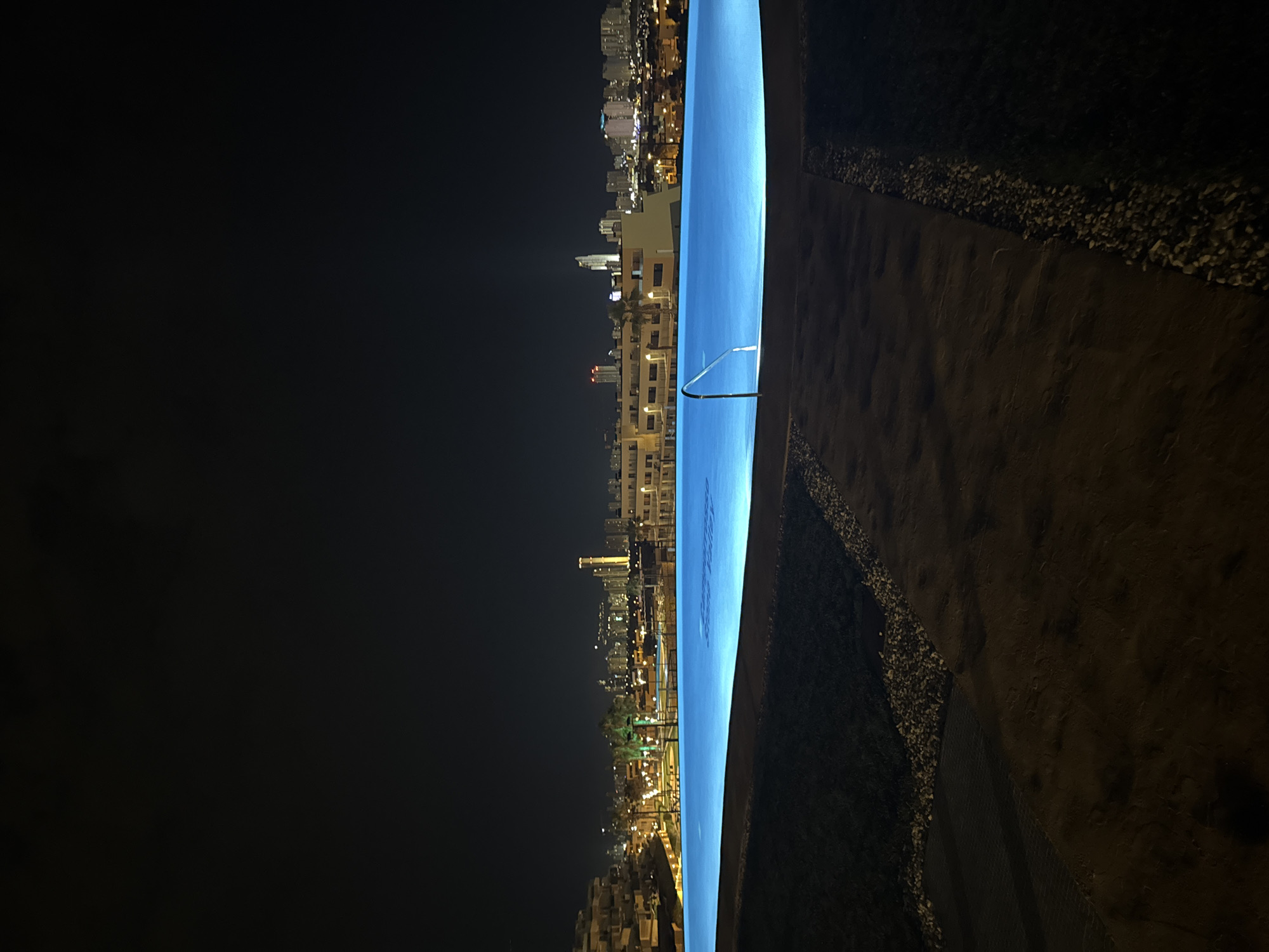 The image shows a night view of an infinity pool with a cityscape in the background. The pool is illuminated with a vibrant blue light, and its surface reflects the lights of the city. The city lights appear as a dense cluster of buildings and skyscrapers. The foreground of the image features the edge of the pool and a dark, textured surface that appears to be a pathway or seating area. There are some scattered small rocks or pebbles along the edge of this surface. The sky is dark and clear, with no visible stars or moon. The architecture of the buildings in the city suggests an urban environment. The overall impression is one of a modern, upscale location, possibly a resort or hotel, offering a scenic view of the city at night. Based on the provided context, this scene is located in Finestrat, Spain. There are no people visible in the image.