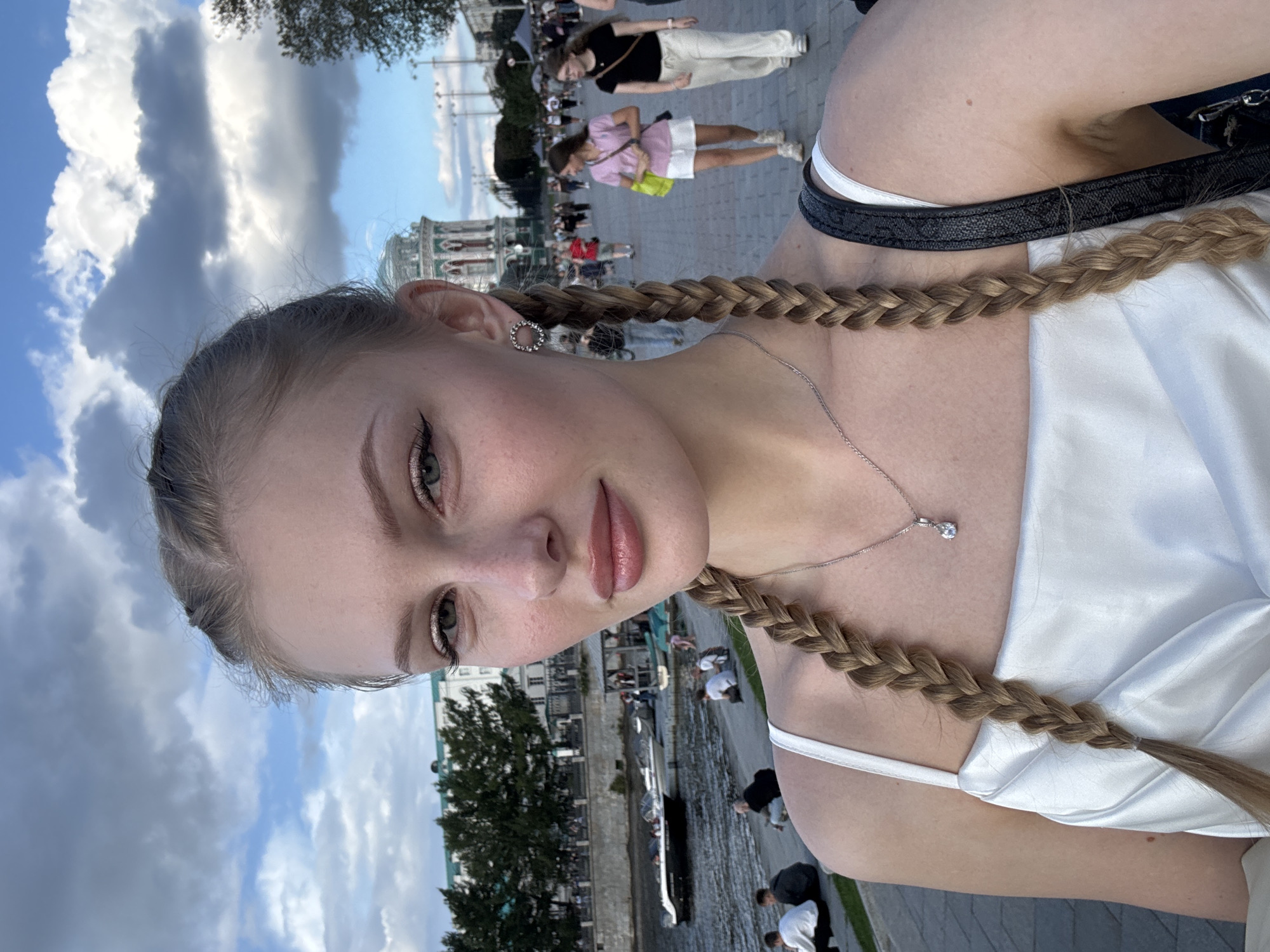 A young woman with blonde hair in two braids is taking a selfie outdoors. She is wearing a white satin top and a delicate silver necklace with a small pendant. She has makeup on, including eyeliner and eyeshadow. In the background, there is a river with boats and people sitting on the embankment. Beyond the river, there are buildings and trees. Further in the background, there are more people walking on a paved area. The sky is cloudy. This scene is identifiable as the embankment of the Iset River in Yekaterinburg, Russia, specifically near the Plotinka (Dam) and the historic buildings of the city.
