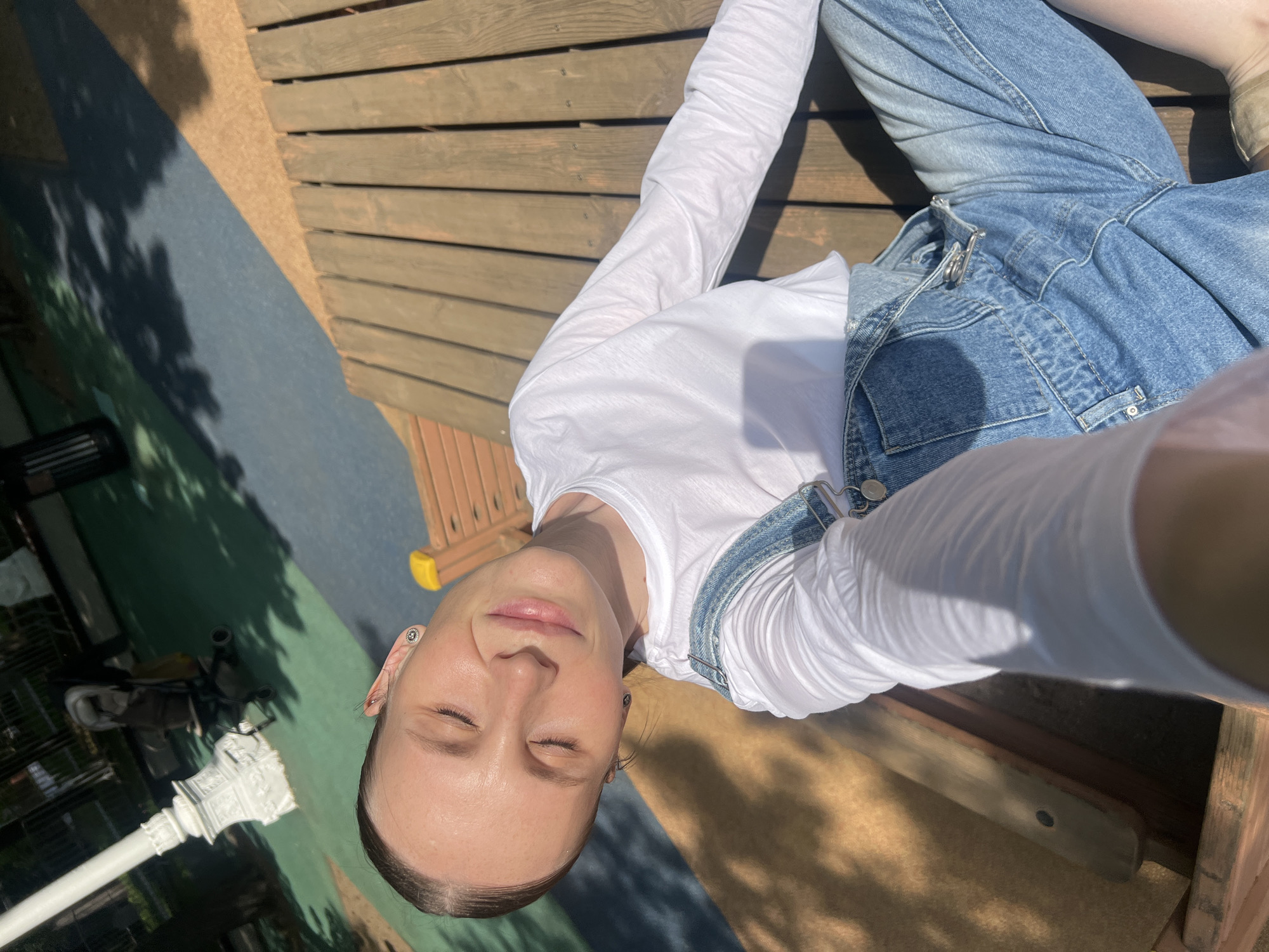 A person is lying on a wooden surface outdoors. The person is wearing blue denim overalls and a long-sleeved white shirt. The ground beneath the wooden surface is a mix of colors, including blue, green, and brown. A white lamppost and a stroller are visible in the background. The scene appears to be in a park or recreation area, with dappled sunlight. Based on the visual details, there is no identifiable information to suggest that this image was taken in Moscow, Russia.