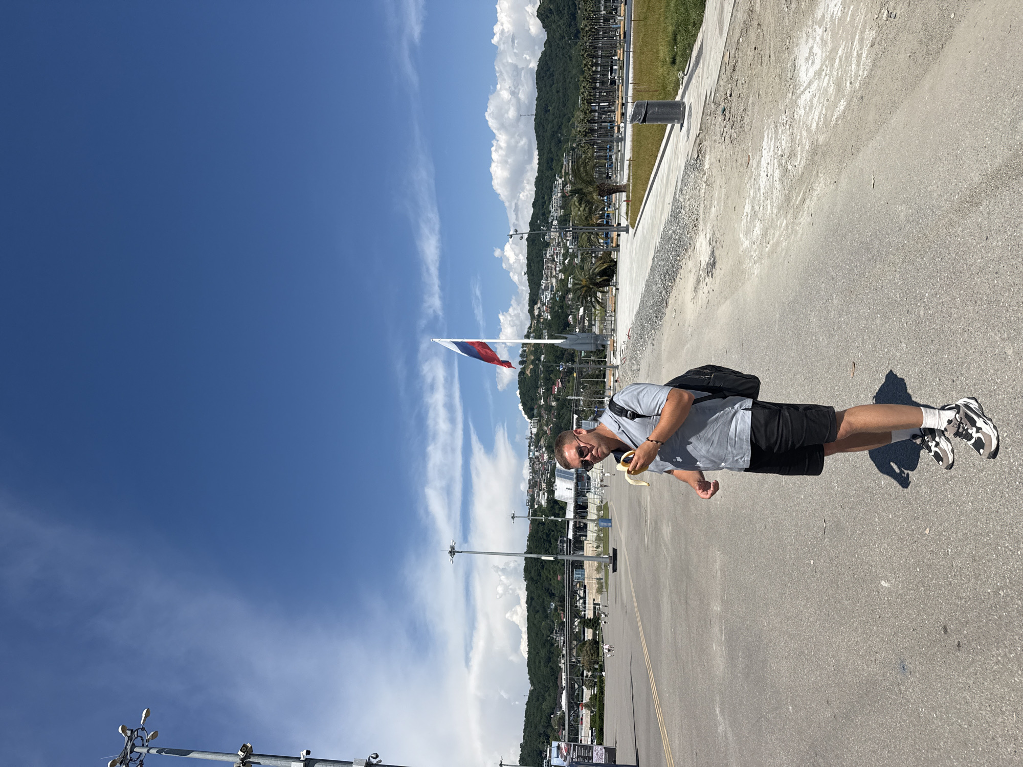 The image depicts a sunlit outdoor scene in Sirius, Russia, likely within the Olympic Park area, characterized by wide paved expanses and modern infrastructure. In the foreground, slightly left of center, a Caucasian male stands facing the camera. He wears dark sunglasses, a light grey short-sleeved polo shirt, black shorts, white socks, and grey, black, and white athletic shoes. A black backpack is slung over his left shoulder. He holds a partially peeled yellow banana in his left hand, appearing to be eating it. His shadow is visible on the asphalt pavement to his right.
The environment consists of a large, grey asphalt surface dominating the foreground and mid-ground, with faint double yellow lines visible on the left. The right edge of the paved area transitions to a strip of gravel and then a neatly maintained grassy section with a dark cylindrical trash receptacle. Tall, multi-fixture streetlights are strategically placed across the paved area, and a large sign with text and graphics is partially visible on the far left.
In the mid-ground, a prominent feature is a tall flagpole from which a large Russian flag (white, blue, red horizontal stripes) flies. Numerous modern buildings with distinct architectural styles are visible, interspersed with rows of palm trees. Densely forested hills and mountains form the distant background, with various residential buildings scattered across their lower slopes under a bright blue sky with scattered cumulus clouds. Several small, indistinct human figures are visible walking in the distance across the paved area.