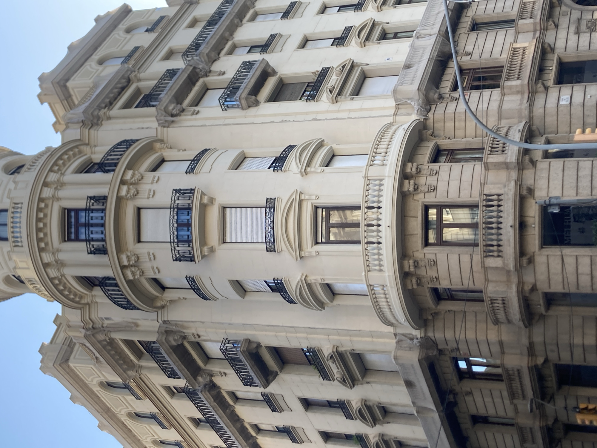 This image depicts the ornate facade of a building in Barcelona, Spain, as seen from a low angle looking upwards. The building features classical architectural elements, including detailed moldings, balconies with wrought-iron railings, and arched windows. The facade is a light, cream-colored stone.
The building appears to be residential or commercial, with numerous windows visible across its height. Some windows have dark frames and appear to be glass, reflecting the sky. Others are covered with blinds or shutters. Decorative balconies jut out from various levels, some with intricate stone carvings. The architectural style suggests a building from the late 19th or early 20th century.
The perspective of the photo is tilted, making the building seem to rise dramatically into the sky. The sky visible is a clear, light blue, indicating a daytime setting with fair weather. The reflection of the building can be seen at the bottom of the image, suggesting the presence of a wet surface, possibly from recent rain or a street cleaning, though the reflection is not perfectly clear. There are no people visible in the image. A traffic light is partially visible on the right side of the frame, providing a subtle cue to an urban environment. No discernible text is present in the image.