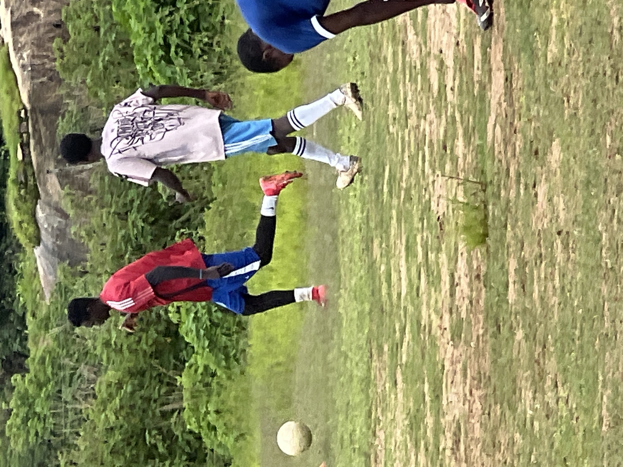 The image depicts a group of young men playing soccer on a dirt and grass field. The setting appears to be outdoors, with lush green vegetation and rocky outcrops in the background. The overall lighting suggests it is daytime, possibly overcast or late afternoon.
In the foreground, a young man wearing a blue jersey and black shorts is partially visible, bent over as if to retrieve or kick a ball. To his left, another young man in a red jersey with white stripes and blue shorts is in motion, his left leg extended forward as if kicking a soccer ball, which is visible in mid-air to his left. He wears black leggings and red soccer cleats. Further to the right, a third young man, seen from the back, wears a light-colored t-shirt with a graphic design and blue shorts, with white socks and cleats. The field is uneven, with patches of dry earth and sparse grass.
The environment suggests a rural or semi-urban outskirts area, possibly in or around Jos, Nigeria, given the natural landscape and the informal setting of a soccer game. The presence of multiple young men engaged in a common activity like soccer points to a community or recreational gathering. The image captures a moment of athletic activity, with focus on the players and the ball.