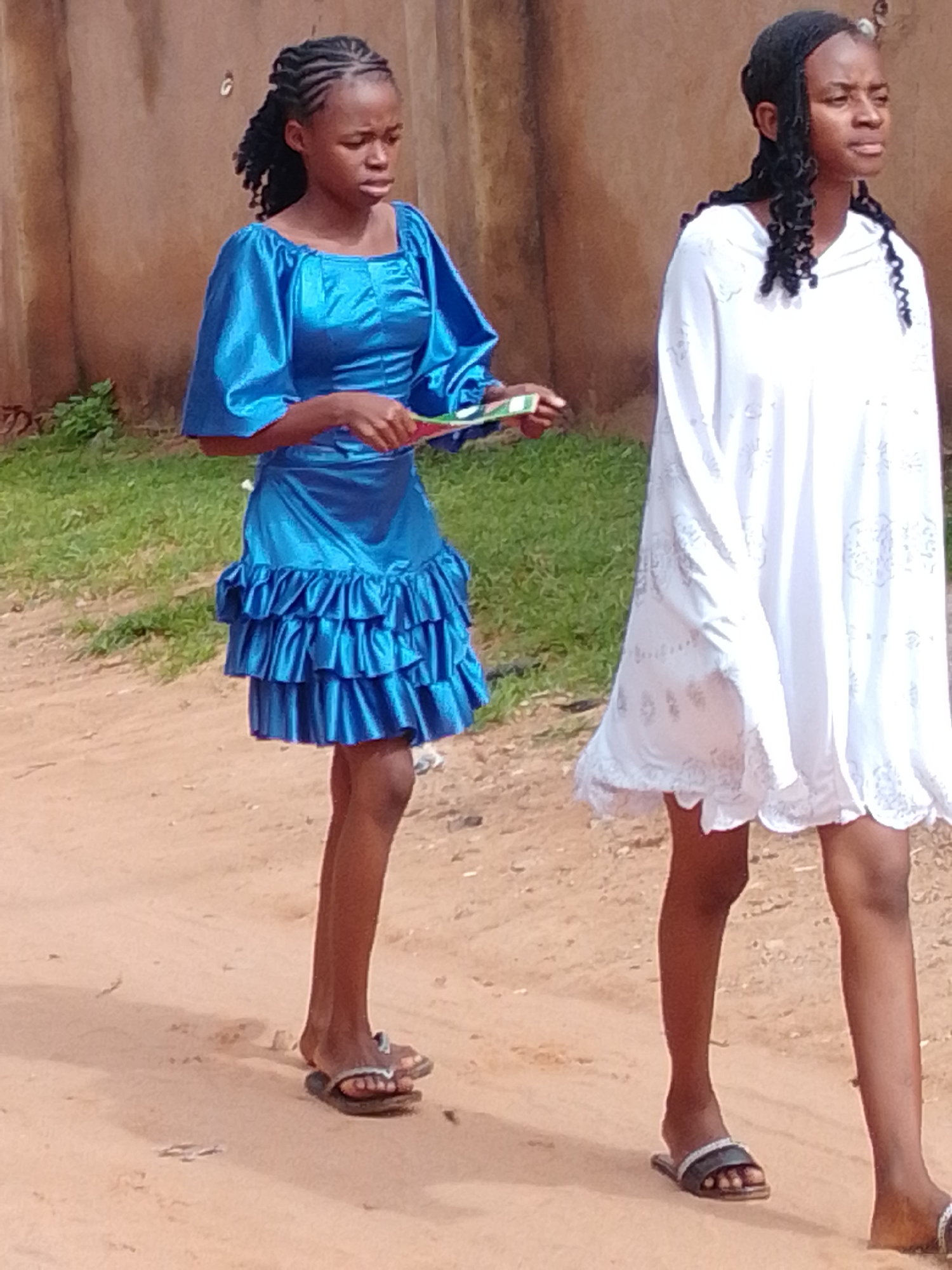 Two young women are walking on a dirt road in front of a tan wall. The woman on the left is wearing a bright blue, satin dress with puffed sleeves and ruffled skirt. She has braided hair and is holding a small, folded object that appears to be a fan or a card. The woman on the right is wearing a long, white, flowing dress with embroidered details on the hem. She has curly hair and is looking to her right. Both women are wearing sandals. The background consists of a rough, tan-colored wall and some green grass. The lighting suggests it is daytime. The scene appears to be in Gombe, Nigeria.
