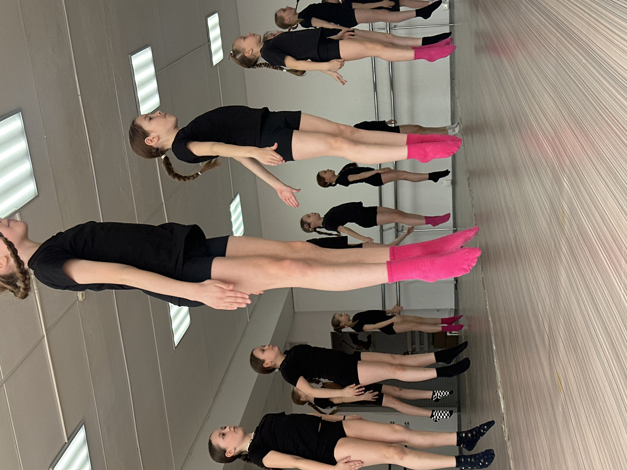 A group of young girls are practicing ballet in a dance studio in Votkinsk, Russia. They are wearing black leotards and shorts with different colored socks. Some have pink socks, while others wear black or patterned socks. They are holding onto a barre for support. The studio has mirrors on one wall, reflecting the dancers. The room is lit by overhead fluorescent lights. A curtain or decorative panel hangs to the right of the barre.
