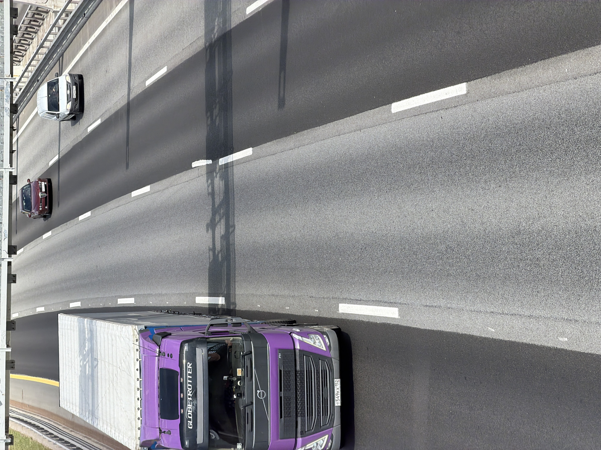 An elevated view captures a highway scene in Saint Petersburg, Russia, on a sunny day. A large purple semi-truck with a white trailer is in the left lane, traveling forward. The truck has the "GLOBETROTTER" branding on its cabin. Behind it, in the same lane, a dark red sedan is also traveling forward. In the right lane, a white van is ahead of the sedan, also moving forward. The highway has multiple lanes divided by white lines, and a metallic overhead structure, possibly a toll booth or signage support, spans across the lanes. Guardrails are visible on the right side of the highway. The road surface appears to be asphalt.