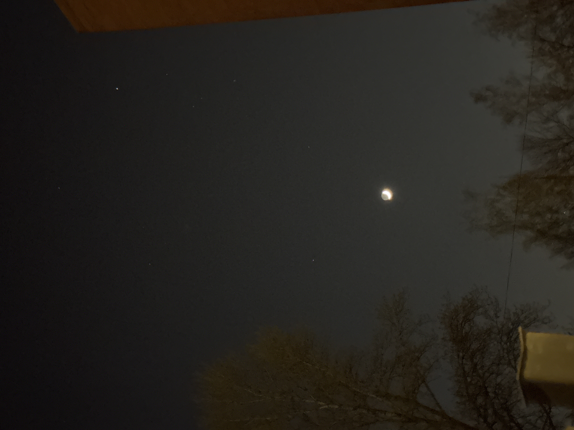 An outdoor, upward-facing shot captures a clear night sky, dominated by a luminous crescent moon positioned off-center towards the upper right. The sky is dark, revealing several faint, pinpoint white lights, likely stars. Silhouettes of bare tree branches, characteristic of deciduous trees in winter or early spring, are visible on the right side and along the bottom edge of the frame, obscuring part of the sky. An architectural element, possibly an overhang or roof, casts a dark shadow across the top left corner. Another light-colored structure, potentially a building or railing, is partially visible in the bottom right corner. The image conveys a serene and crisp night scene, with no discernible activity or people. There is no visible text. The time of day is clearly night, and the sky appears clear.