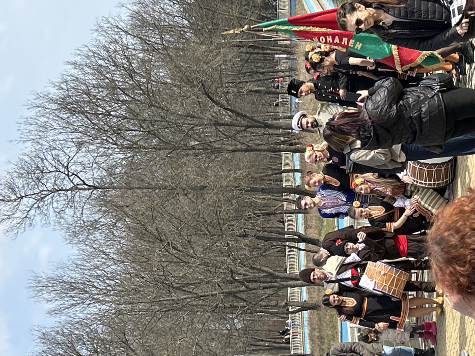 The image captures an outdoor gathering in Borisova Gradina park, Sofia, Bulgaria. Approximately 15 individuals, primarily adults with a few children, are positioned in the mid-ground, posing for a photograph. The majority are dressed in elaborate Bulgarian folk costumes featuring embroidered shirts, vests, skirts, and patterned sashes. Noteworthy attire includes a male in a light military-style uniform with a white peaked cap, another male in a dark uniform with a black fur hat, and several females with floral hair adornments. Two large cylindrical wooden drums (tupans) are visible, one held by a man on the left and another partially obscured, with a third resting on the ground to the right. A flagpole on the far right displays a partially visible flag with green, white, and red stripes, likely the Bulgarian national flag, and Cyrillic text. In the immediate foreground, the backs of spectators' heads are visible, with one woman actively photographing the costumed group using a smartphone. The background is dominated by numerous bare deciduous trees under a clear blue sky, suggesting late winter or early spring. A light-blue lined water basin or canal, flanked by a white metal railing, extends horizontally across the mid-ground behind the primary group, with other park visitors visible distantly beyond it.