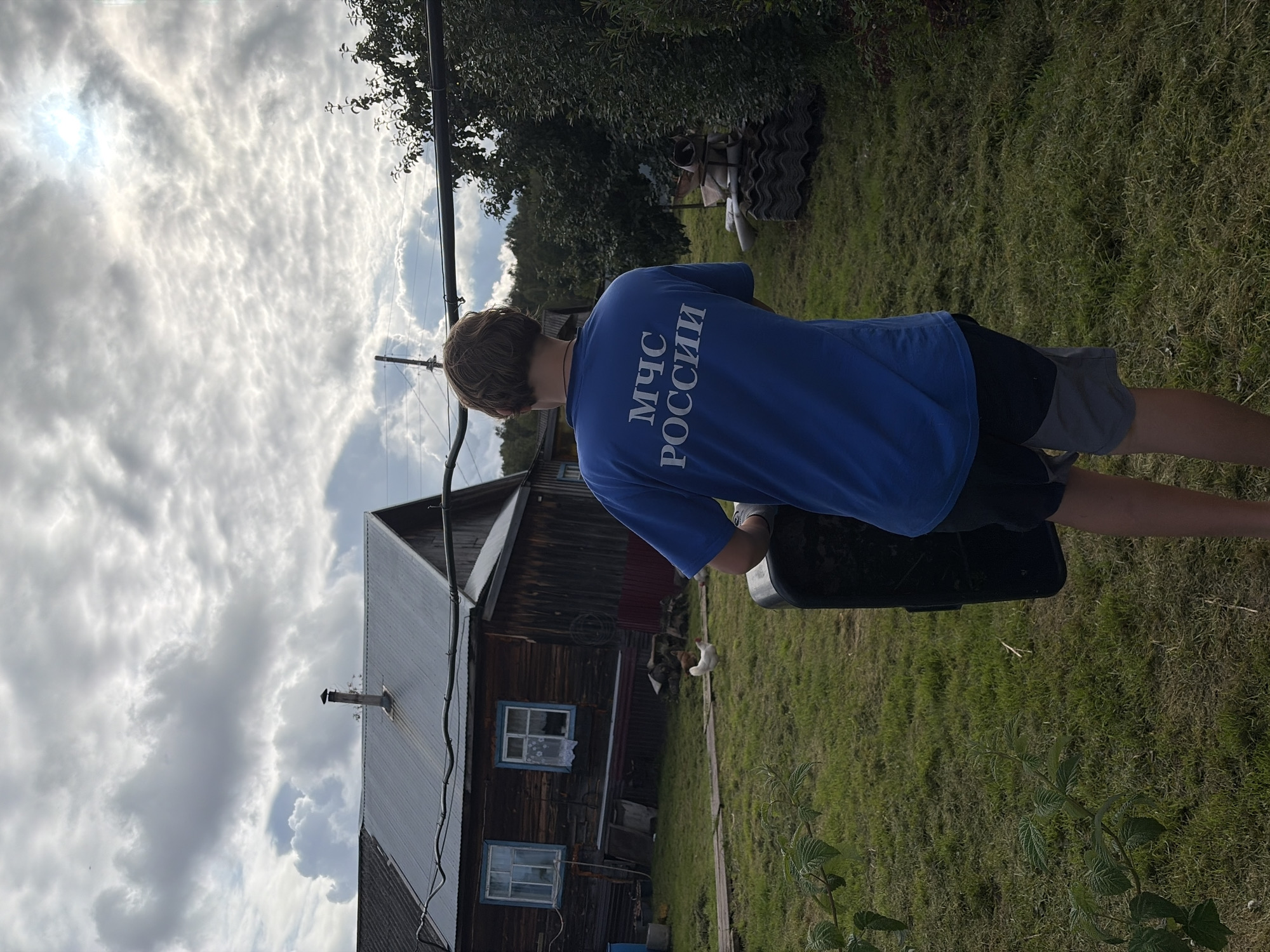 An individual wearing a blue t-shirt with "МЧС РОССИИ" (EMERCOM of Russia) printed on the back is standing in a yard, facing away from the camera. They are holding a black, rectangular object, possibly a suitcase or a shield, in their right hand. The person is wearing dark shorts. The yard is covered in green grass, with some patches of mown grass visible. In the background, there is a wooden house with a metal roof and two windows. A chimney is visible on the roof. There are some chickens in the yard. The environment appears to be rural, possibly in a village. The sky is cloudy with patches of sunlight breaking through. The image is taken outdoors. The location is identified as Узяр, Russia.