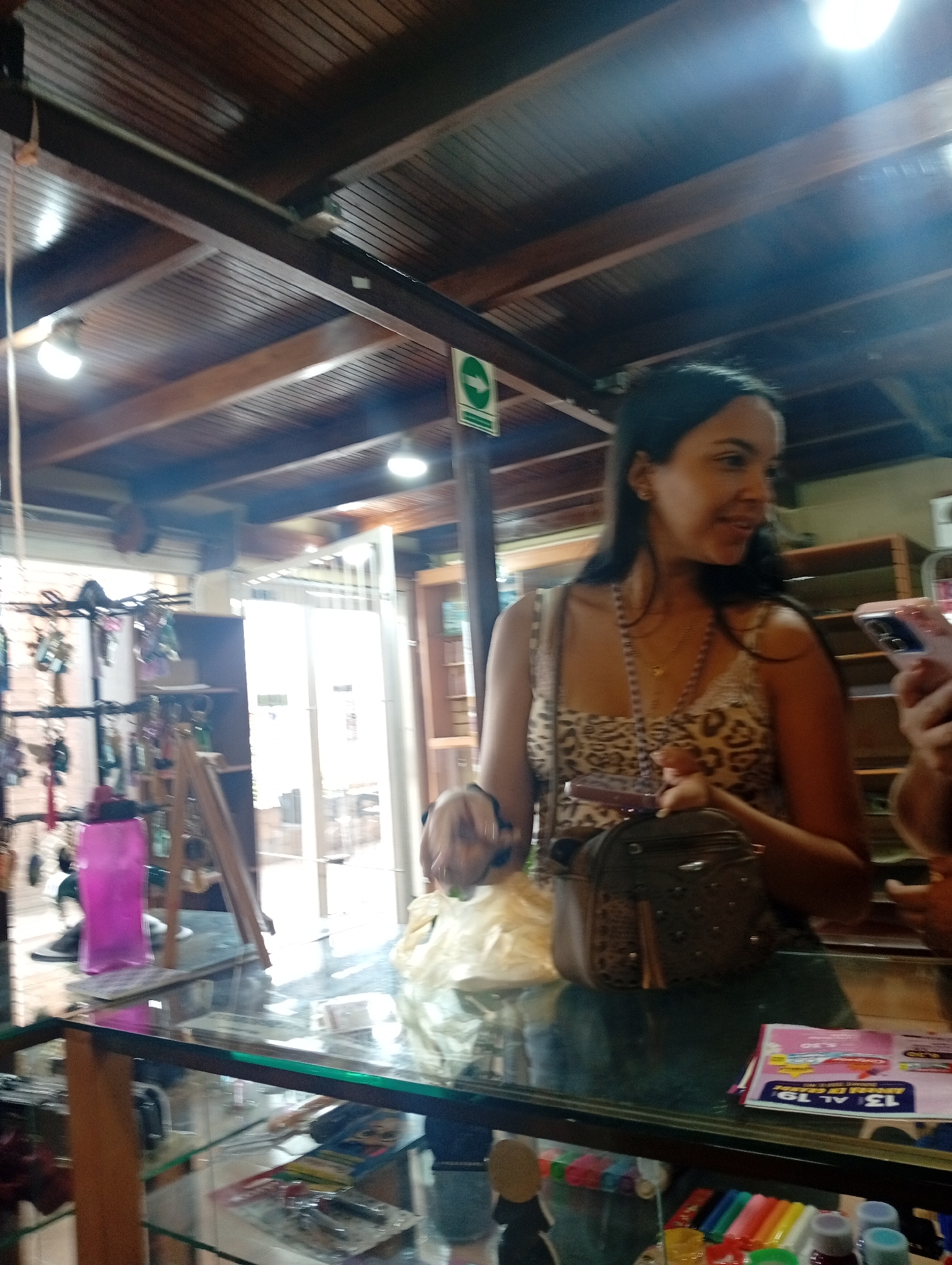 The image depicts an indoor scene within a shop or store, likely in Juan Griego, Venezuela. The setting is characterized by a prominent wooden ceiling featuring dark beams and lighter wooden panels, illuminated by several circular overhead lights.
The main subject is a woman, visible from the chest up, standing behind a glass counter. She has dark hair and is wearing a leopard-print sleeveless top and a long, beaded necklace with a small pendant. Her expression is pleasant and engaged, with a slight smile as she looks towards her right. One hand rests near a dark brown or grey handbag with tassel and stud details, placed on the counter. Part of another person's hand, holding a smartphone with a pink case, is visible on the far right.
The glass counter appears to be part of a display case. On its surface, an open magazine or flyer is visible, displaying text such as "13 AL 19" and "BS. 6,99". Below the counter, inside the display, various small items like colorful markers, pens, and small bottles are arranged. To the left, behind the counter, a vibrant pink water bottle stands beside a display rack laden with numerous keychains and other small decorative items.
In the background, a brightly lit doorway or large window suggests it is daytime. A green square sign with a white arrow pointing right, typical of an exit or directional sign, is mounted on a wooden beam near the ceiling. Additional shelves are visible further back. The overall impression is that of a retail environment, where the woman is either shopping or engaging in a transaction at the counter.
