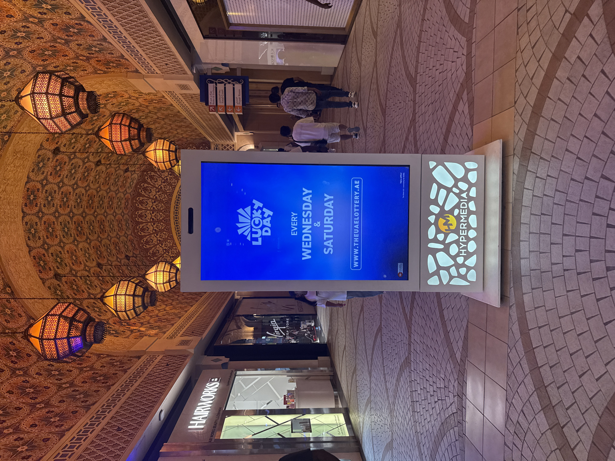 The image captures an indoor walkway in Dubai, United Arab Emirates, characterized by traditional Arabian architectural elements. Ornate lamps hang from the intricately patterned ceiling, casting a warm glow. The walls and ceiling are adorned with detailed mosaics and geometric designs. Shops line the walkway, including "HAIRWORKS" and "Virgin MEGA STORE," identifiable by their signage. In the foreground, a large digital advertising screen displays a promotion for "LUCKY DAY," announcing it occurs "EVERY WEDNESDAY & SATURDAY" with the website "WWW.THEUAELOTTERY.AE." Below the screen, the "HYPERMEDIA" logo is visible. Several people are walking through the corridor, some are seen from behind, appearing to be shoppers. The floor is paved with interlocking stones in a patterned arrangement. The overall atmosphere suggests a modern shopping mall with a distinct cultural aesthetic.