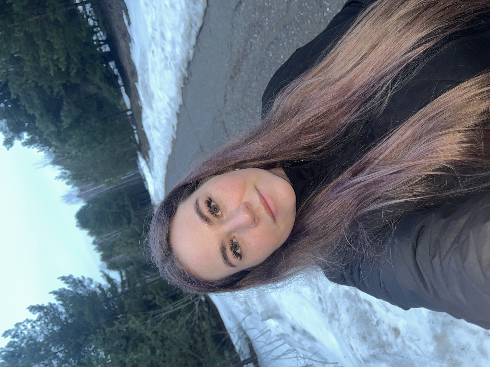 A woman takes a selfie outdoors. She has long, light brown hair with purple streaks, fair skin, and is wearing a black jacket. The background includes a paved road, patches of snow, and coniferous trees, suggesting a park or wooded area. The location is likely the city of Chaikovsky, Russia.