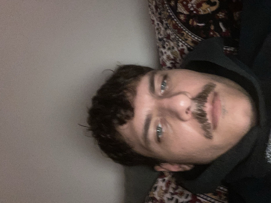 A selfie depicts a man with curly brown hair and striking light blue eyes. He sports a distinctive, somewhat unusual mustache. He wears a dark-colored hoodie. The background is split between a plain, light-colored wall on one side and a patterned rug or blanket on the other. The image is taken from a slightly upward angle, giving a unique perspective on the subject.