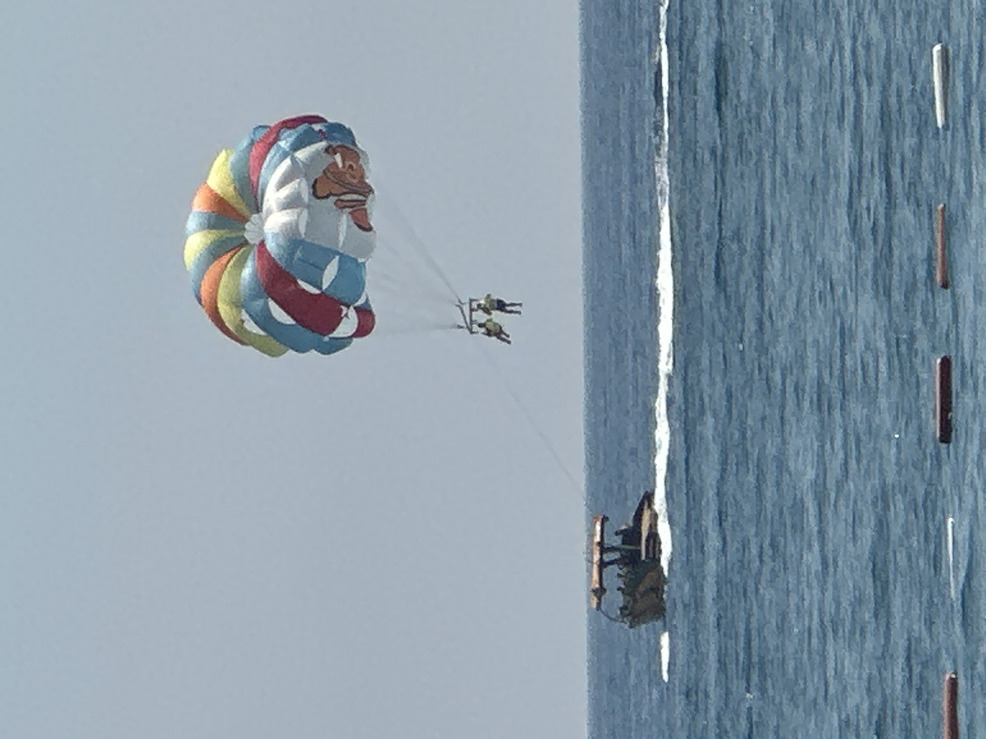 In the city of Boreti, Montenegro, a parasailing activity is taking place on a clear, overcast day. A colorful parasail, featuring a character resembling Nemo from Finding Nemo, is suspended in the sky. Two individuals are attached to the parasail, appearing to be enjoying the ride. Below, a boat with a canopy and several passengers is speeding across the blue water, creating a white wake. The sea is choppy, with visible waves. The image captures a moment of recreational activity in an outdoor, marine environment.